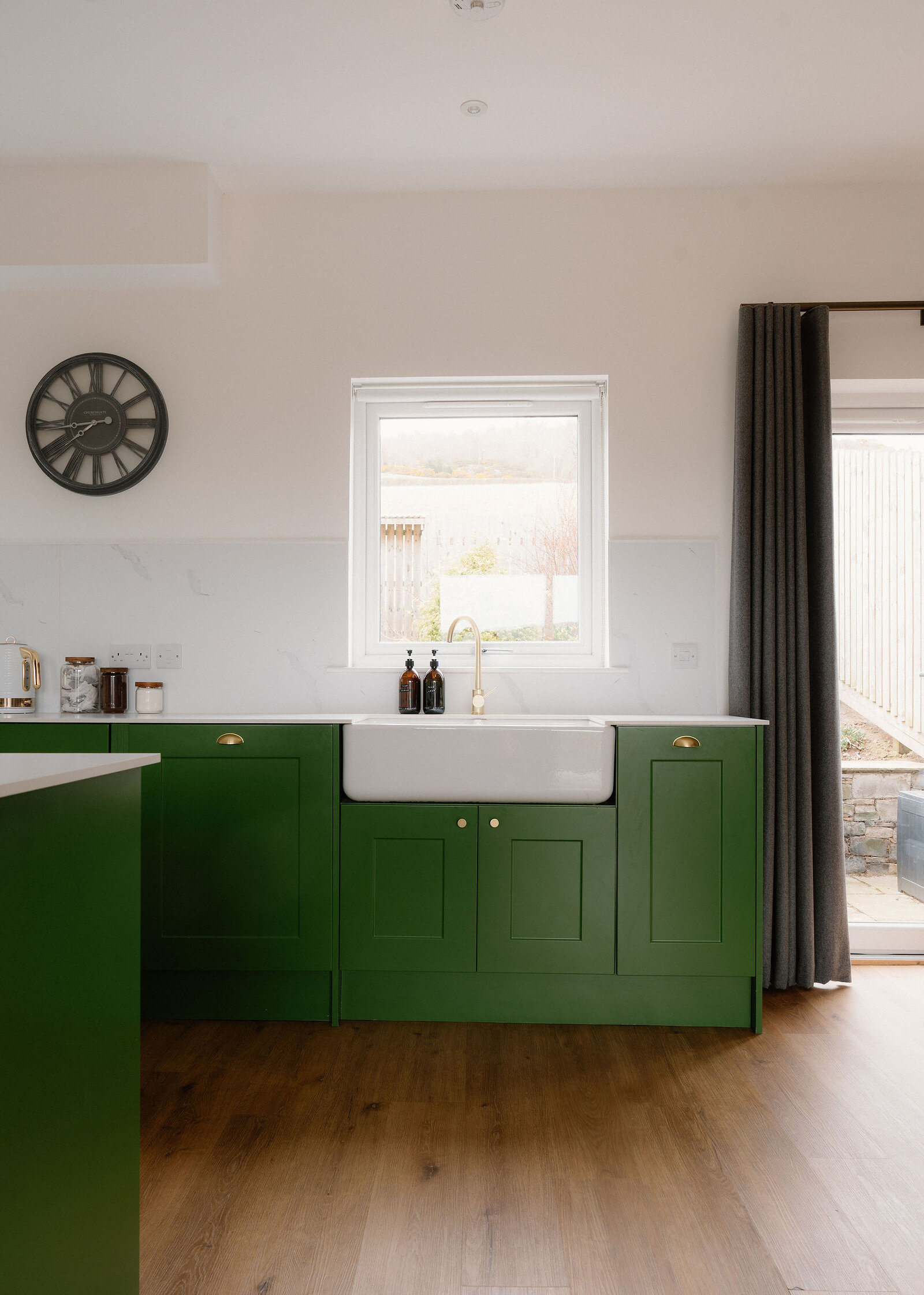 A green kitchen with a Belfast sink and wall clock has jars set on the counter beside a curtained glass door.