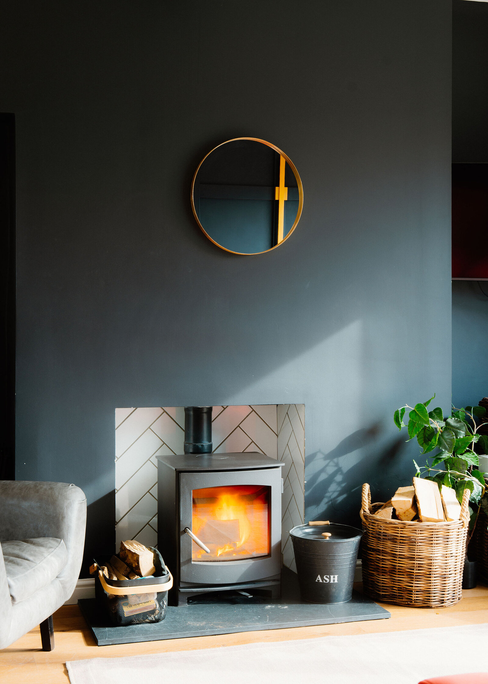 A wood-burning stove burns in a herringbone-tiled recess below a round brass mirror, a log carrier, ash bucket and plant beside it.