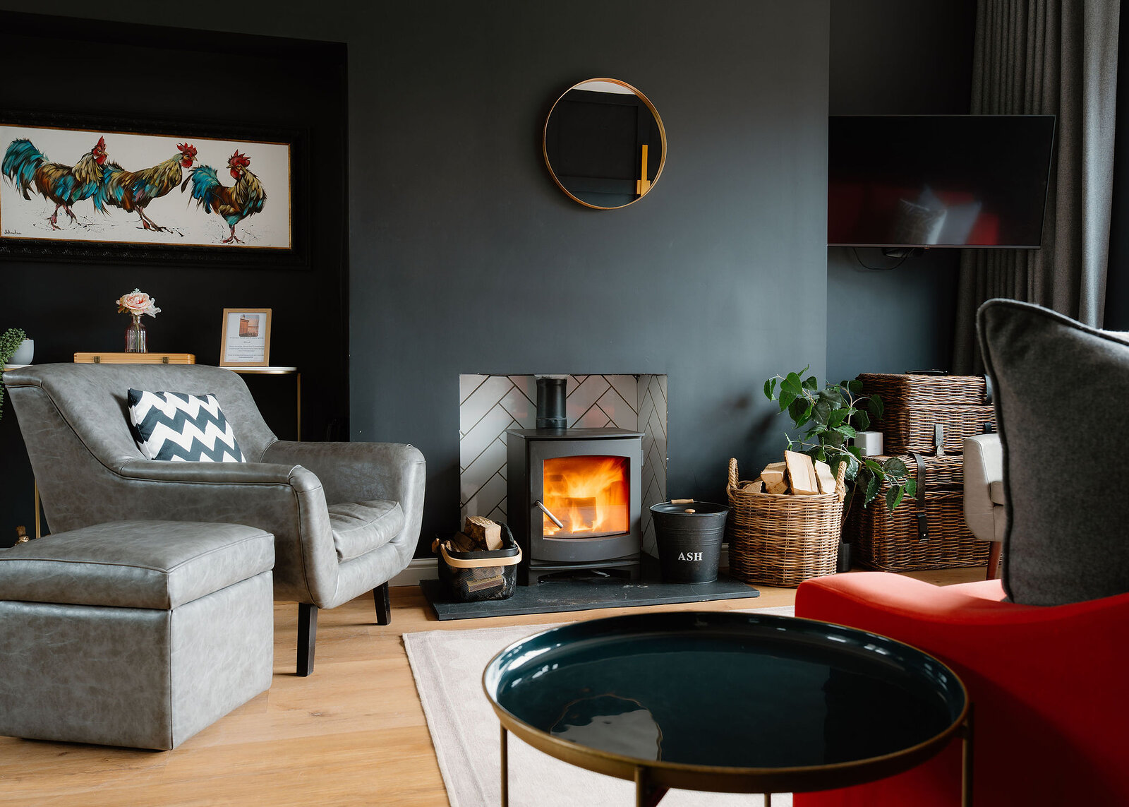 A grey armchair with a striped cushion sits before a lit wood-burning stove, a framed print of chickens and a red sofa corner nearby.