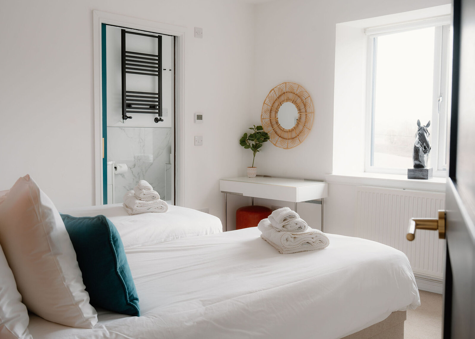Twin beds with folded towels and a teal cushion sit before a dressing table and rattan mirror, a black towel rail through the bathroom door.