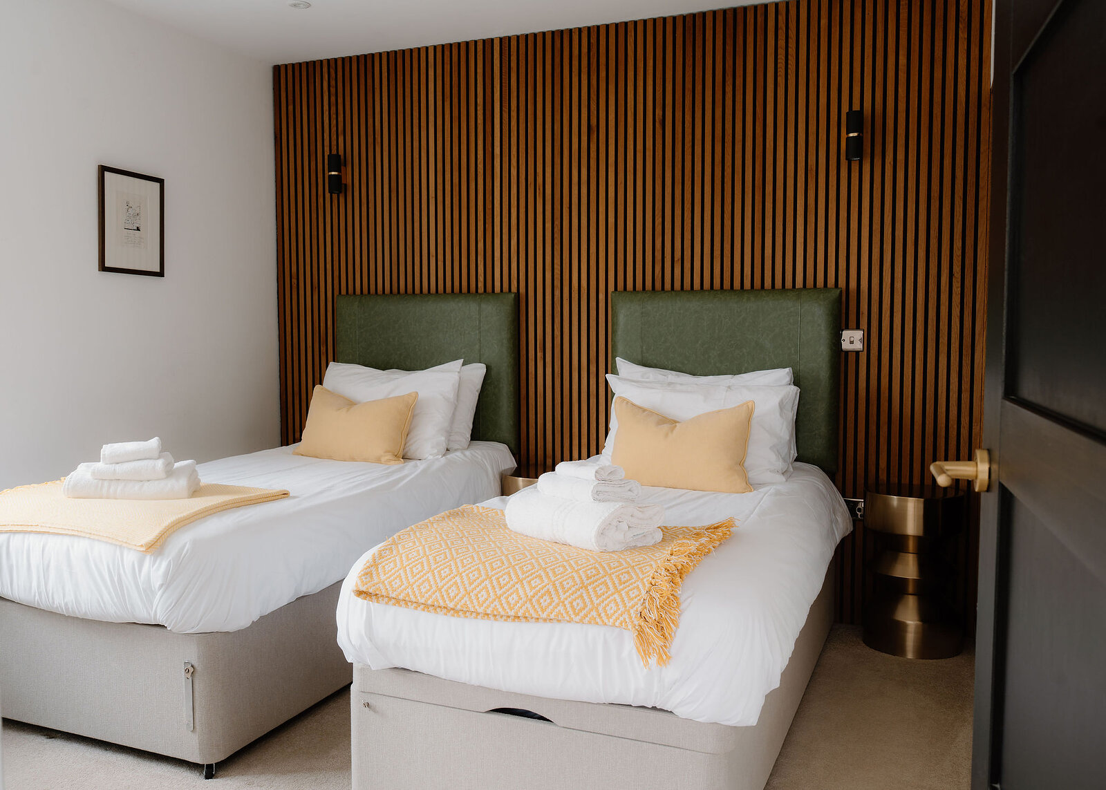 Twin beds with green leather headboards and yellow throws stand against a wall of slatted oak panelling, with a brass side table.