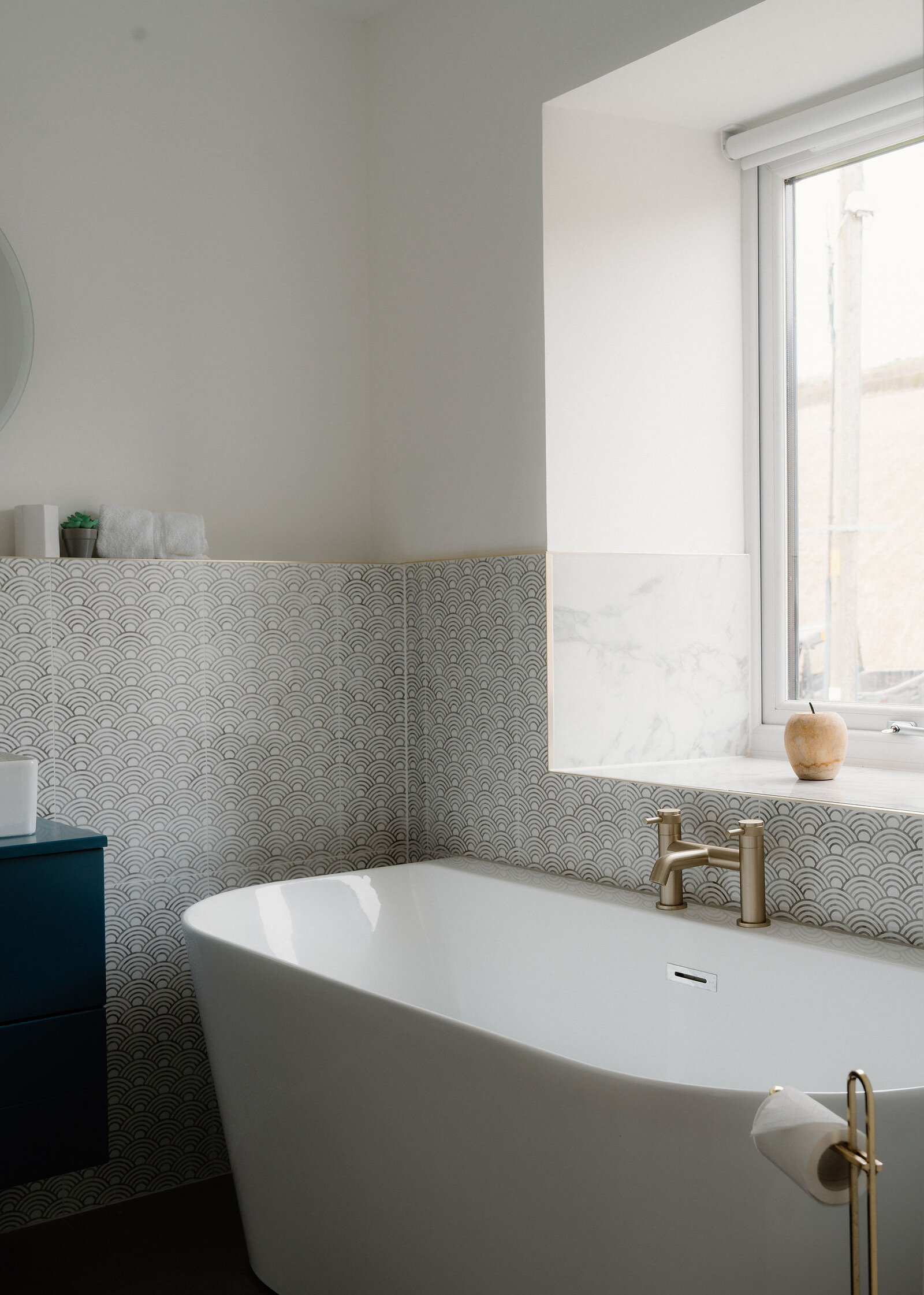 A freestanding bath sits beneath a window with a patterned tiled surround, an apple resting on the sill beside the gold tap.