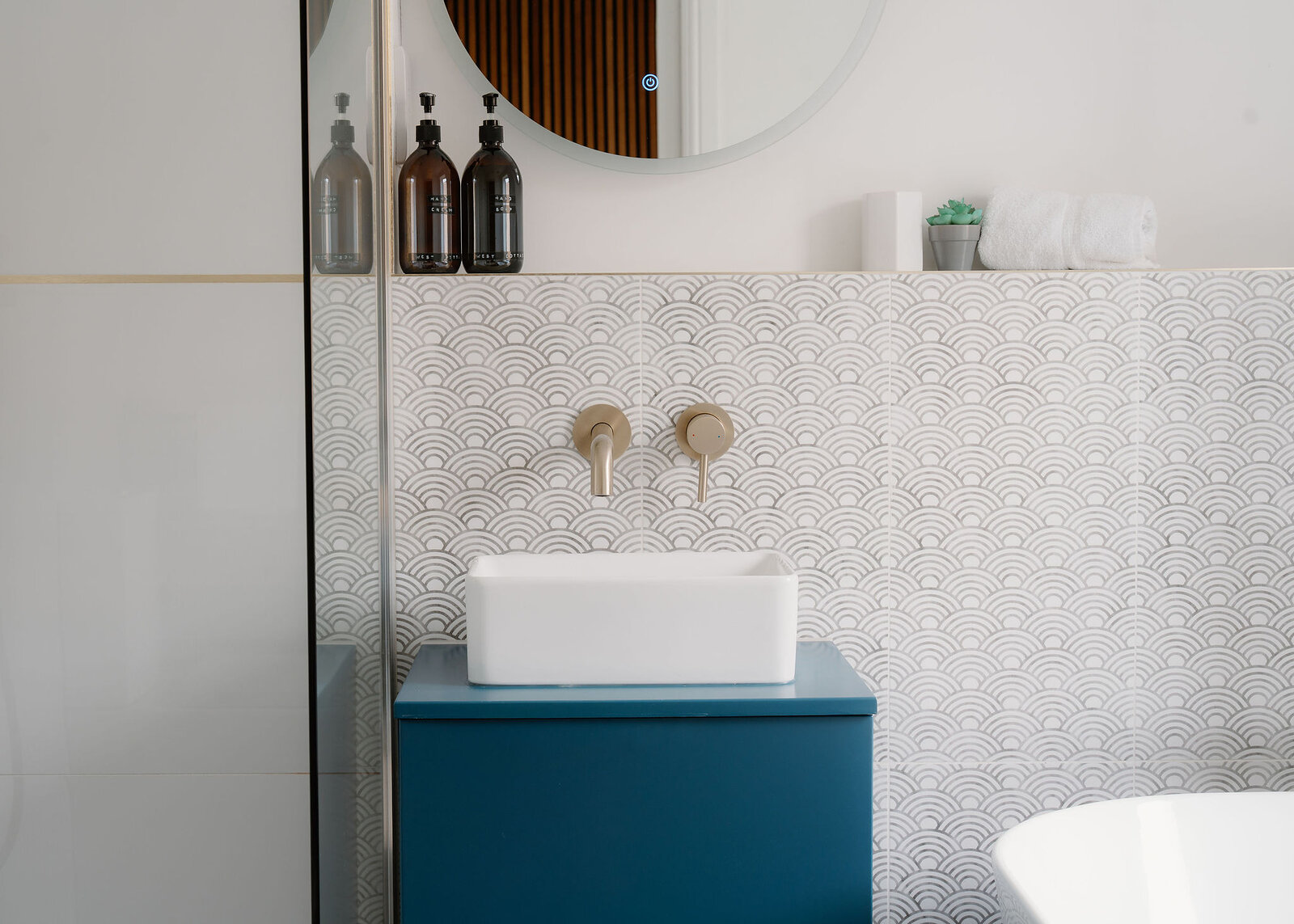 A teal vanity unit with a white basin sits below gold wall-mounted taps and a round mirror, toiletry bottles on a shelf against patterned tiles.