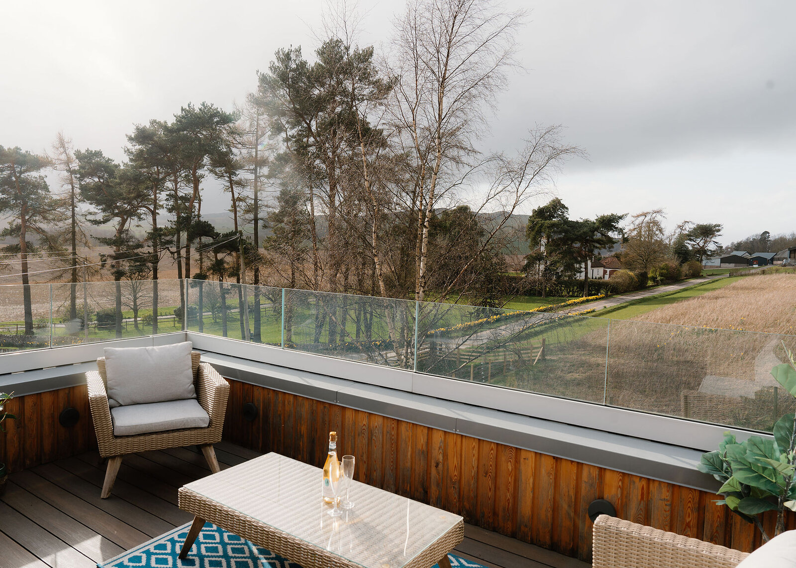 A wicker armchair sits on a decked balcony behind a glass balustrade, a coffee table with a bottle and glass in the foreground and trees and fields beyond.