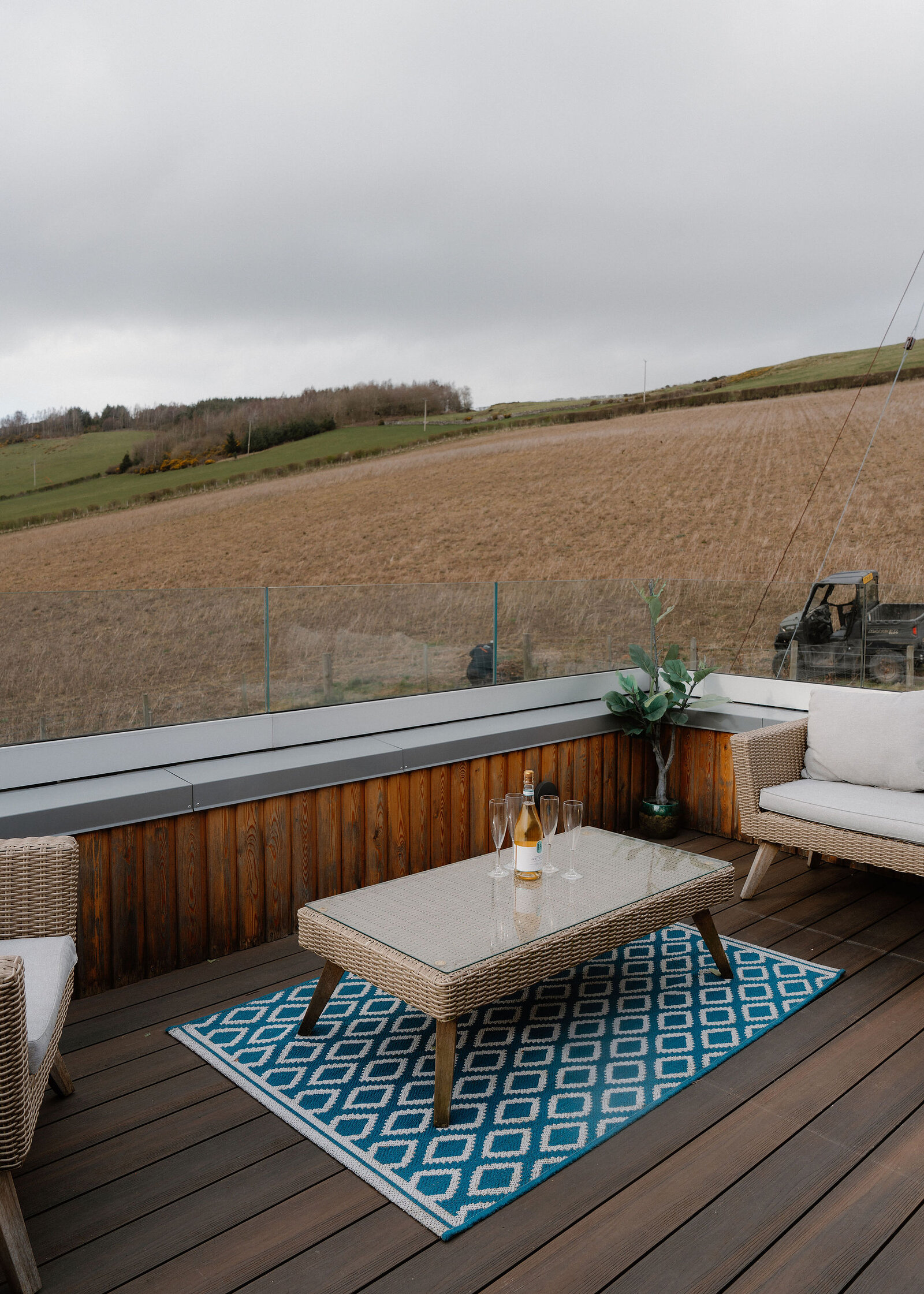Wicker armchairs and a sofa surround a glass-topped table with a bottle and flutes on a decked balcony above a stubble field.