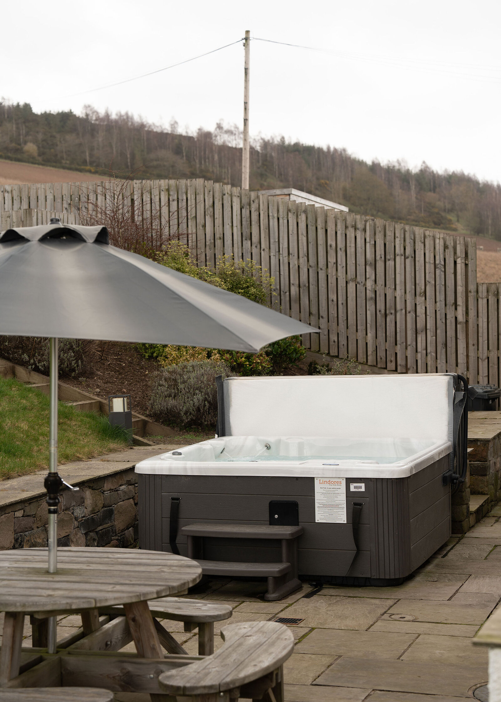 A hot tub with its lid raised stands on a paved terrace behind a picnic table and grey parasol, below a wooded hill.