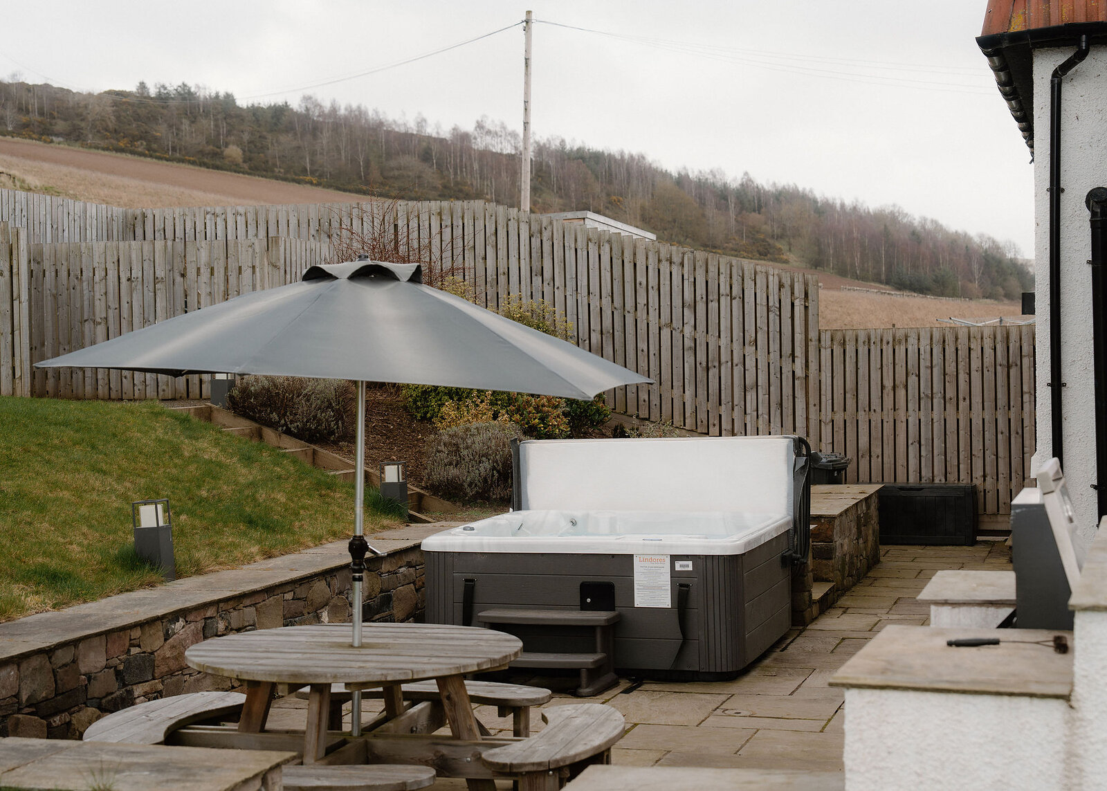 The paved garden terrace seen whole, with the hot tub, a picnic table under a parasol and a built-in barbecue against the house.