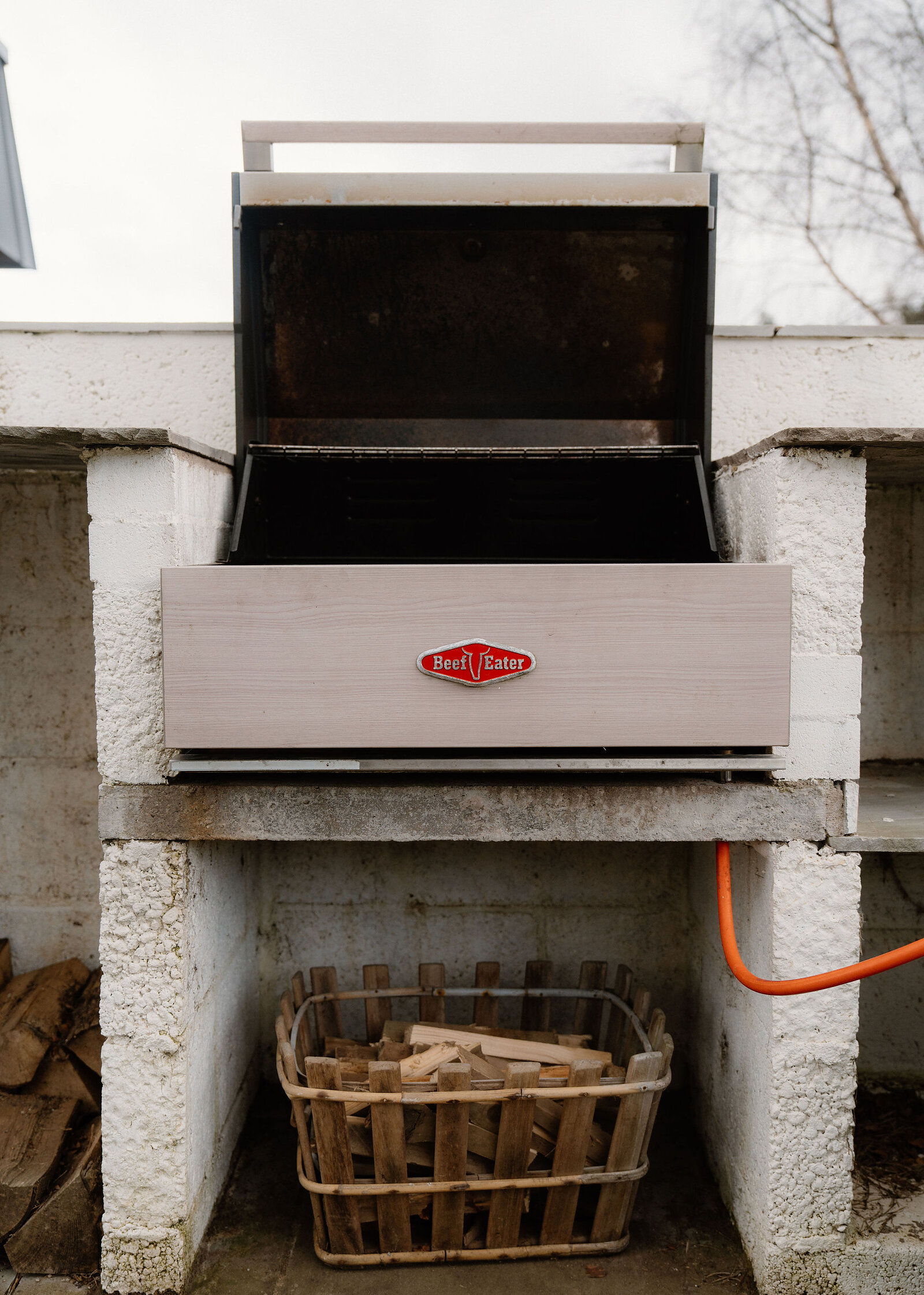 A built-in barbecue grill stands open on a whitewashed block surround, a basket of logs stored in the space beneath.