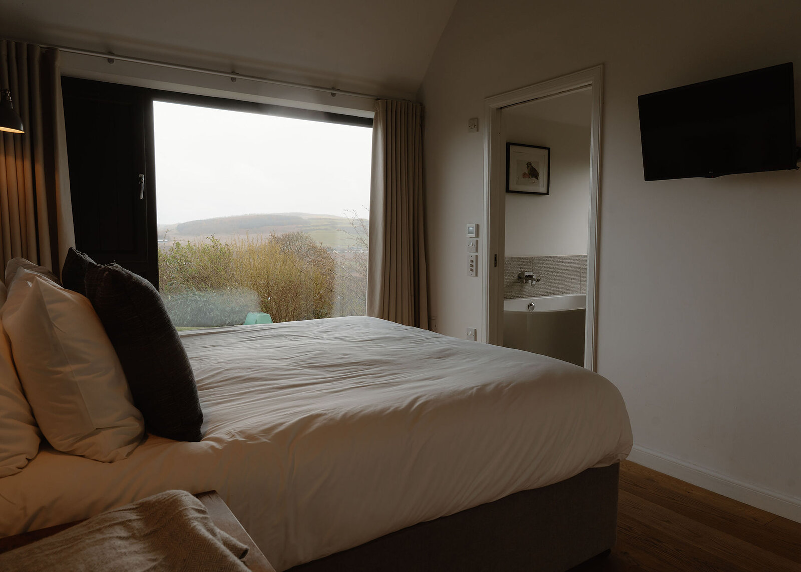 A bed with grey cushions faces a tall window onto shrubs and a hazy hill, with an en-suite door open at right.