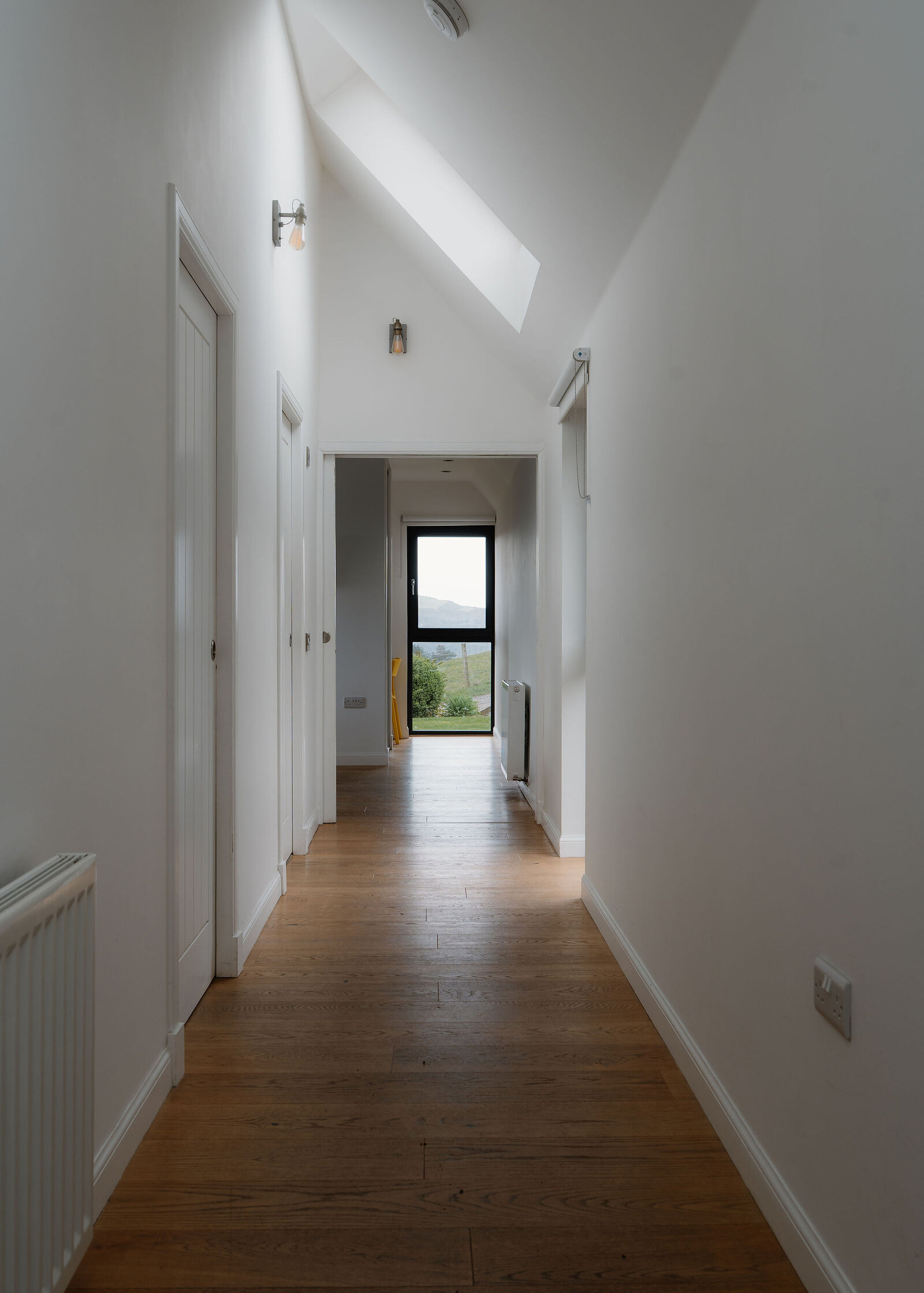 A hallway with a vaulted ceiling and a skylight leads past wall lights to a window at the far end.
