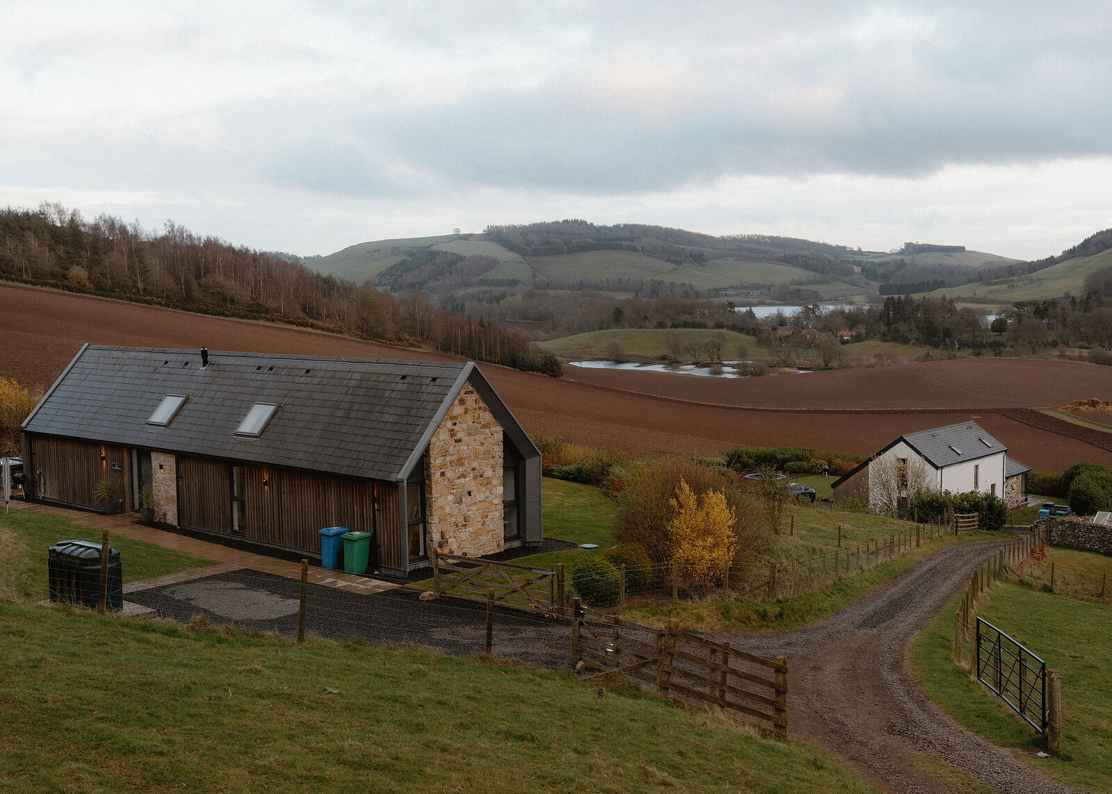 The timber and stone house with a farm track curving past a white house, towards a loch below ploughed fields and green hills.