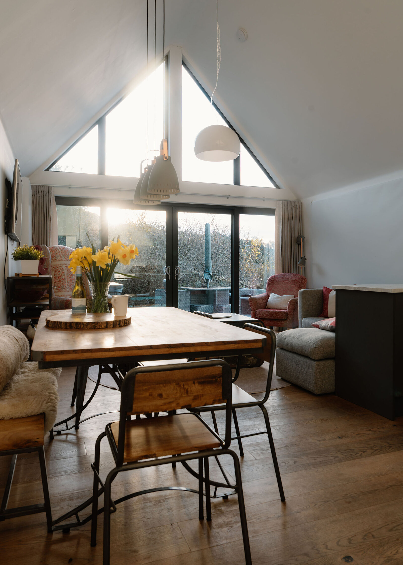 A wooden dining table with a vase of daffodils sits beneath pendant lights in a room with a pitched glazed gable end, a patterned armchair and glass doors to the garden.