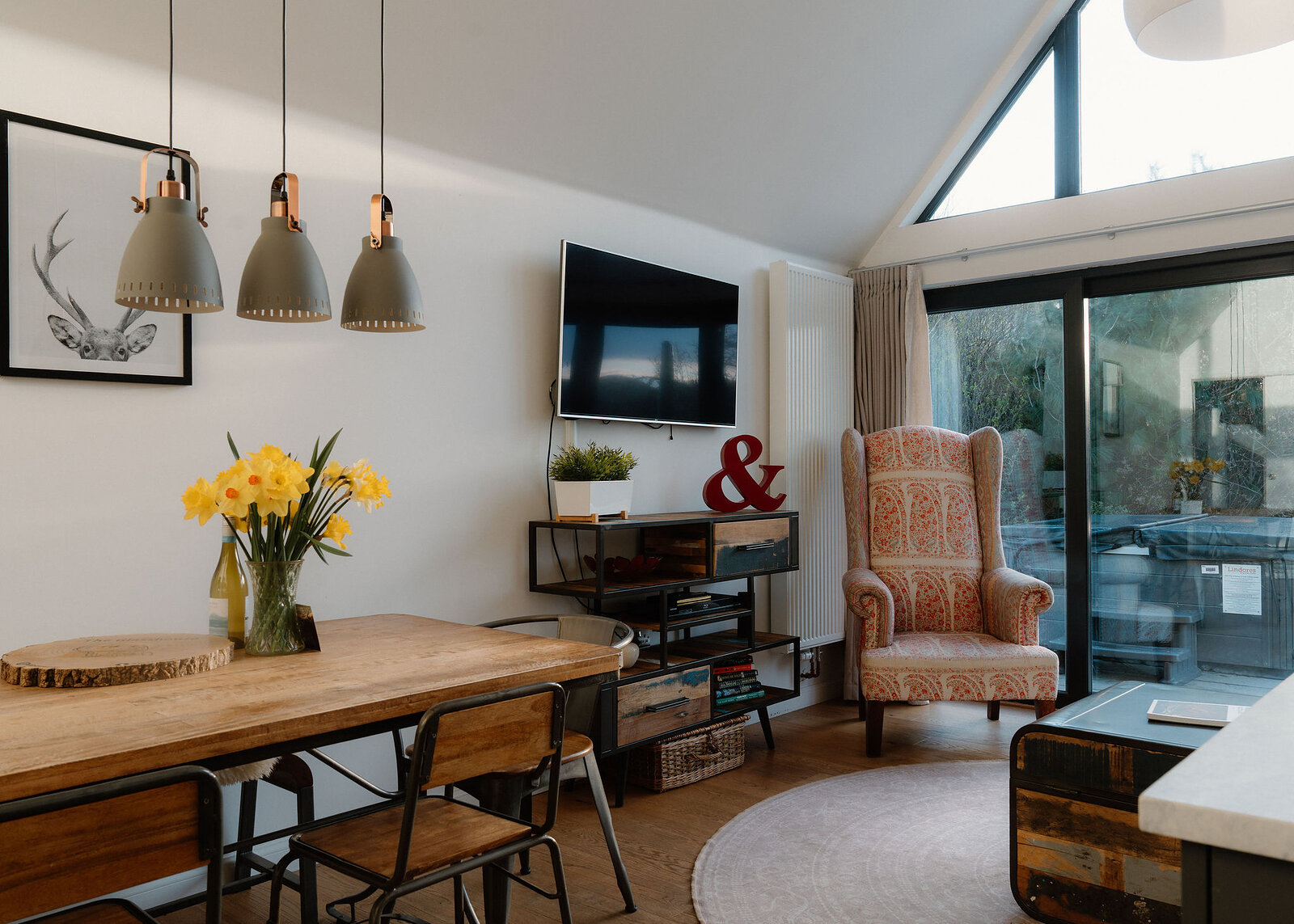 Three pendant lights hang above a wooden dining table with daffodils, a patterned wingback armchair and a wall-mounted television nearby.