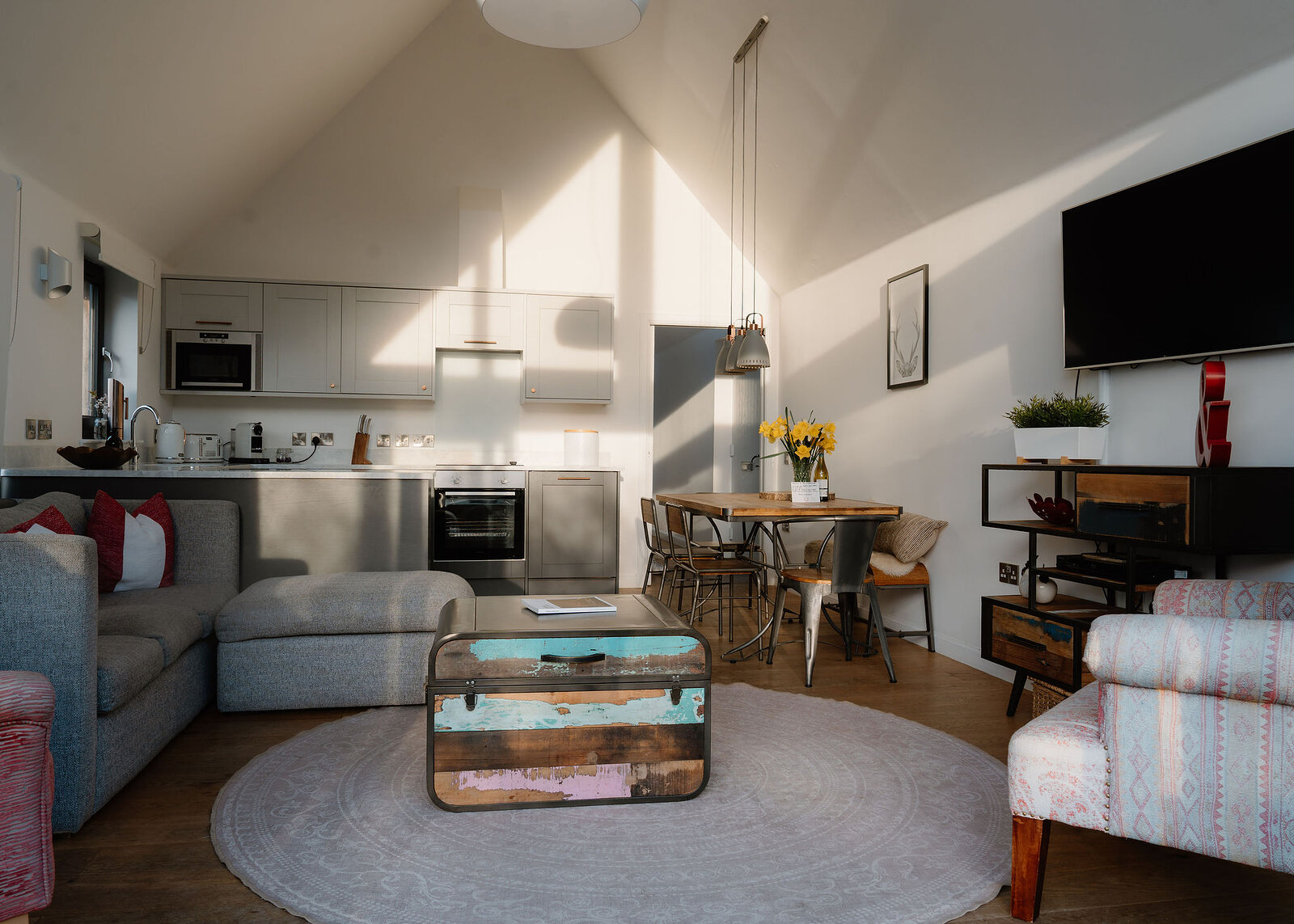 A grey corner sofa surrounds a painted wooden trunk used as a coffee table, a kitchen, dining table and patterned armchair beyond.