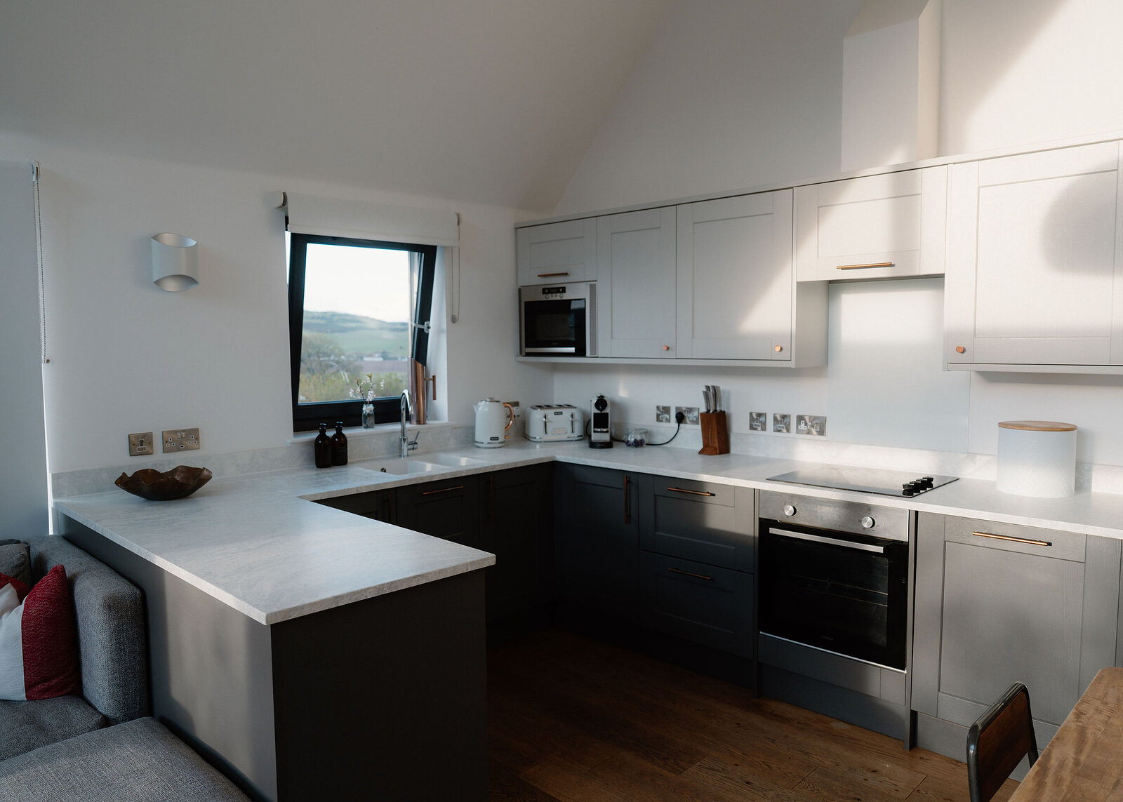 A U-shaped kitchen with dark grey cabinetry and white worktops includes an oven, hob and microwave, a window above the sink looking out to fields.