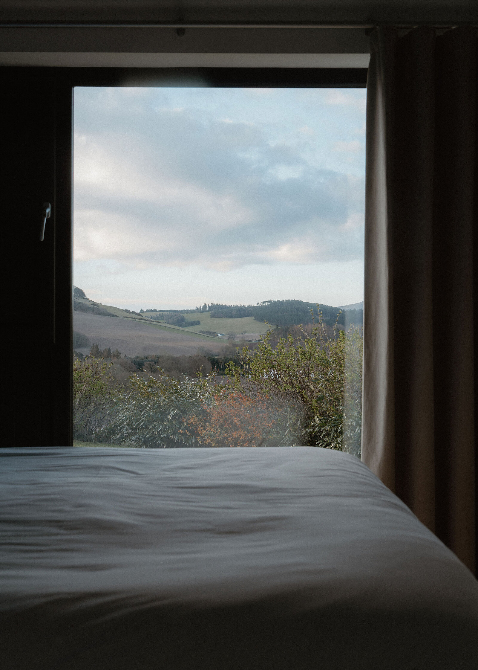 The same bedroom window taken closer and centred, framing shrubs, ploughed fields and wooded hills under a blue-grey sky with broken cloud.