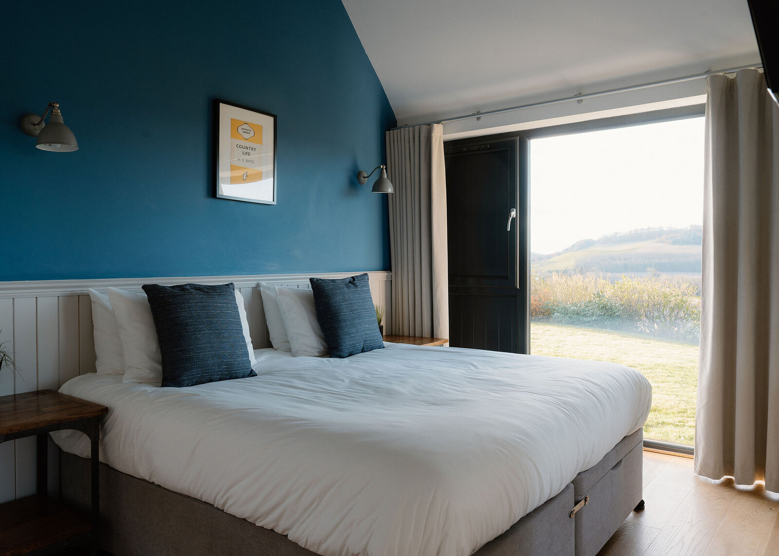 A bed with white bedding and navy cushions sits against a deep teal wall hung with a small framed print, a glazed door open to a view of fields beyond.