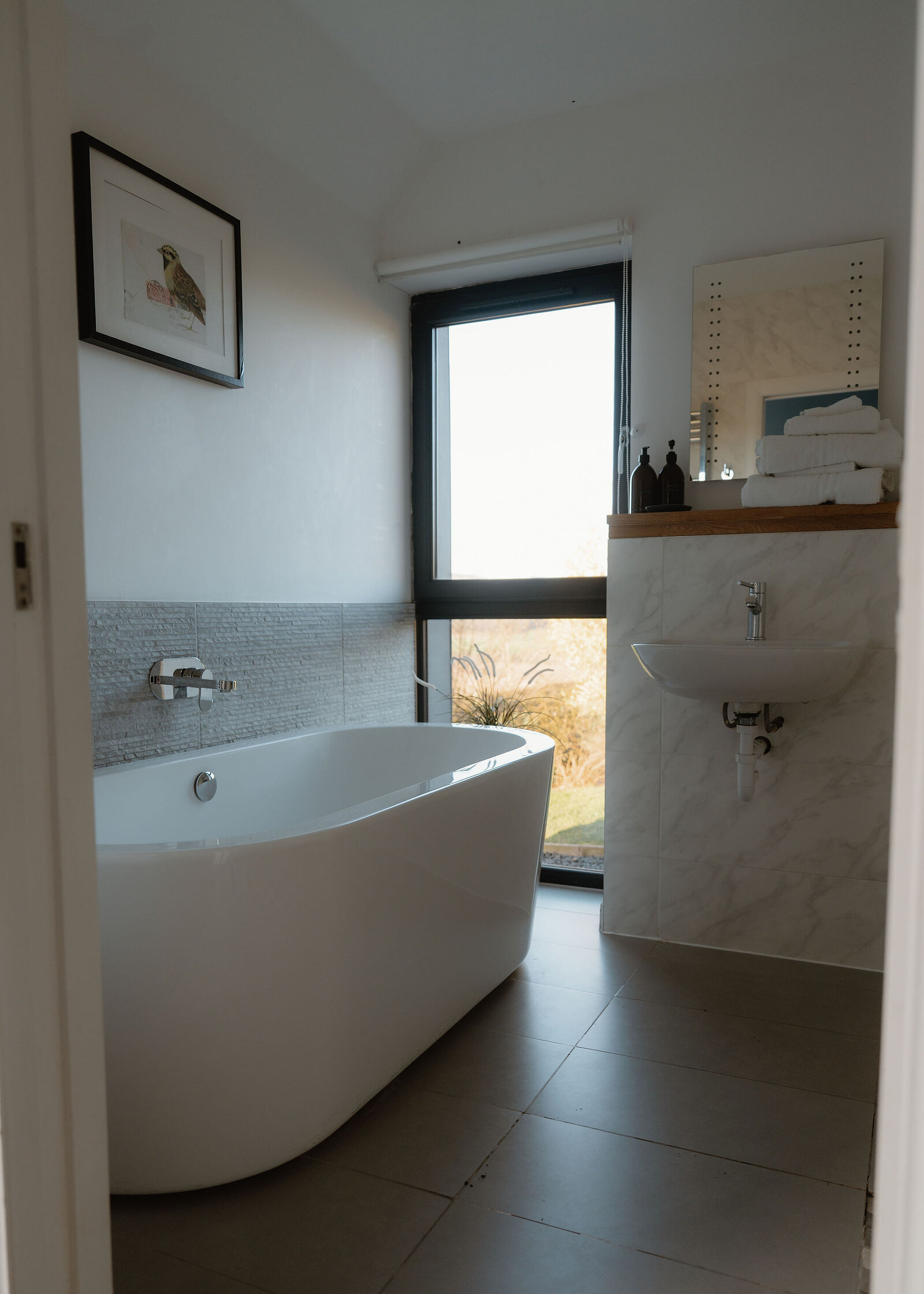A freestanding bath sits below a bird print beside a tall window and a basin, with folded towels and bottles on a timber shelf.
