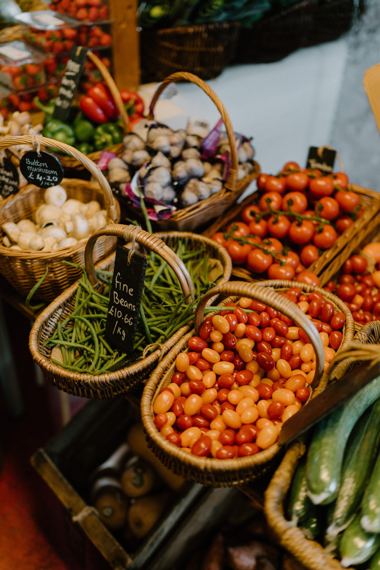 Wicker baskets of cherry tomatoes, green beans, mushrooms and garlic sit on a market stall, each labelled with a handwritten price.
