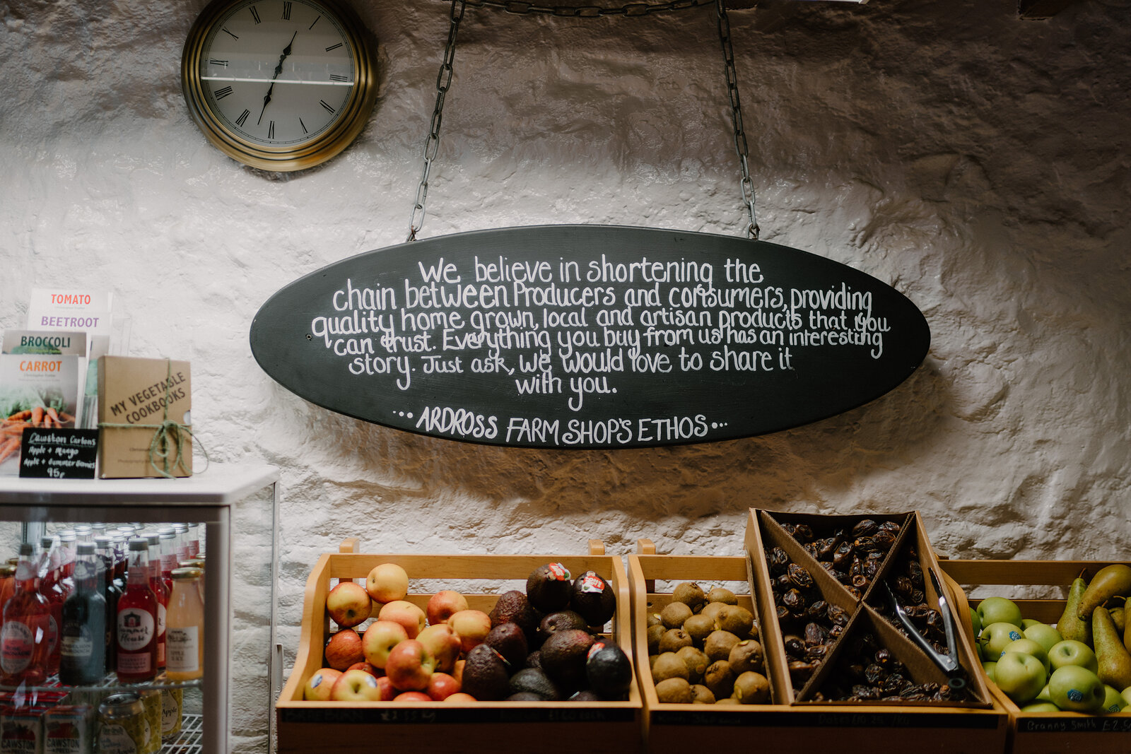 A hand-lettered oval chalkboard describing a farm shop's ethos of shortening the chain between producers and consumers hangs above crates of apples, chestnuts and pears, a wall clock mounted beside it.