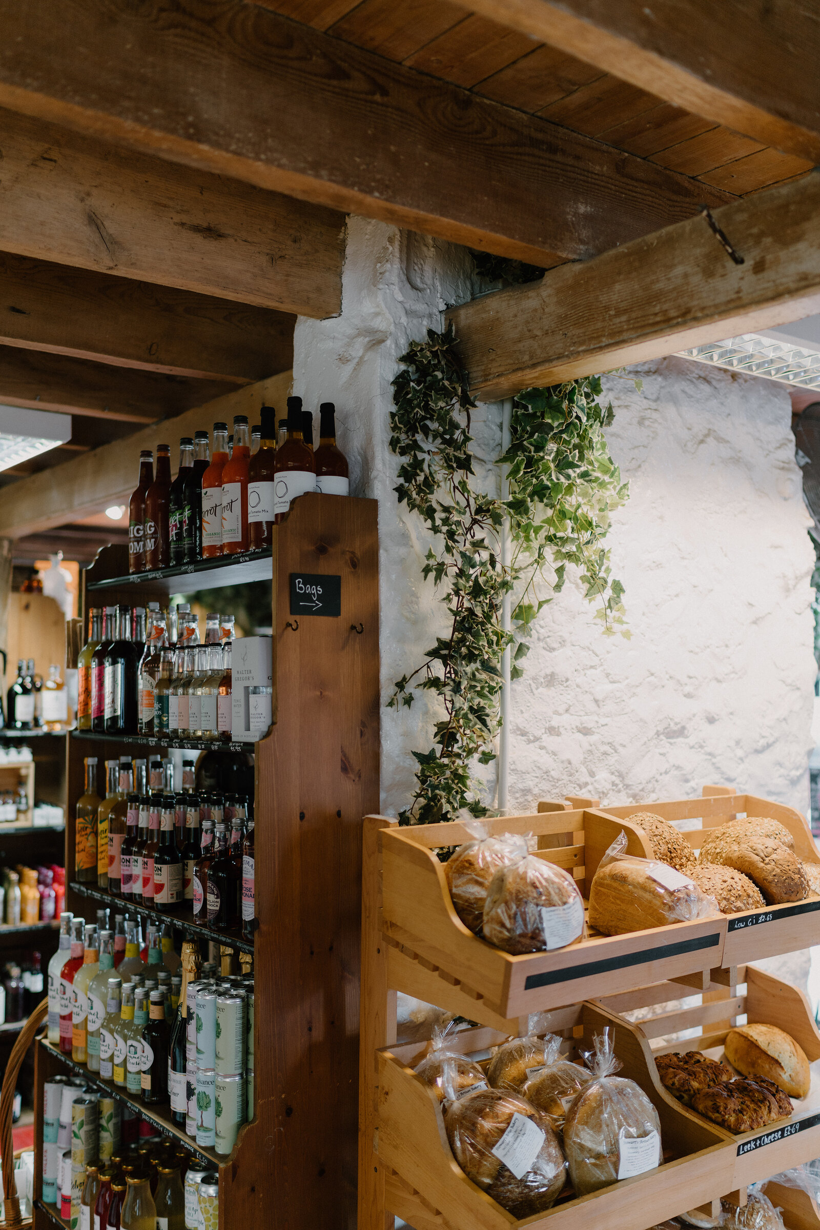 A wooden shelf of bottled drinks stands beside baskets of bread loaves, ivy trailing down the whitewashed stone wall behind.