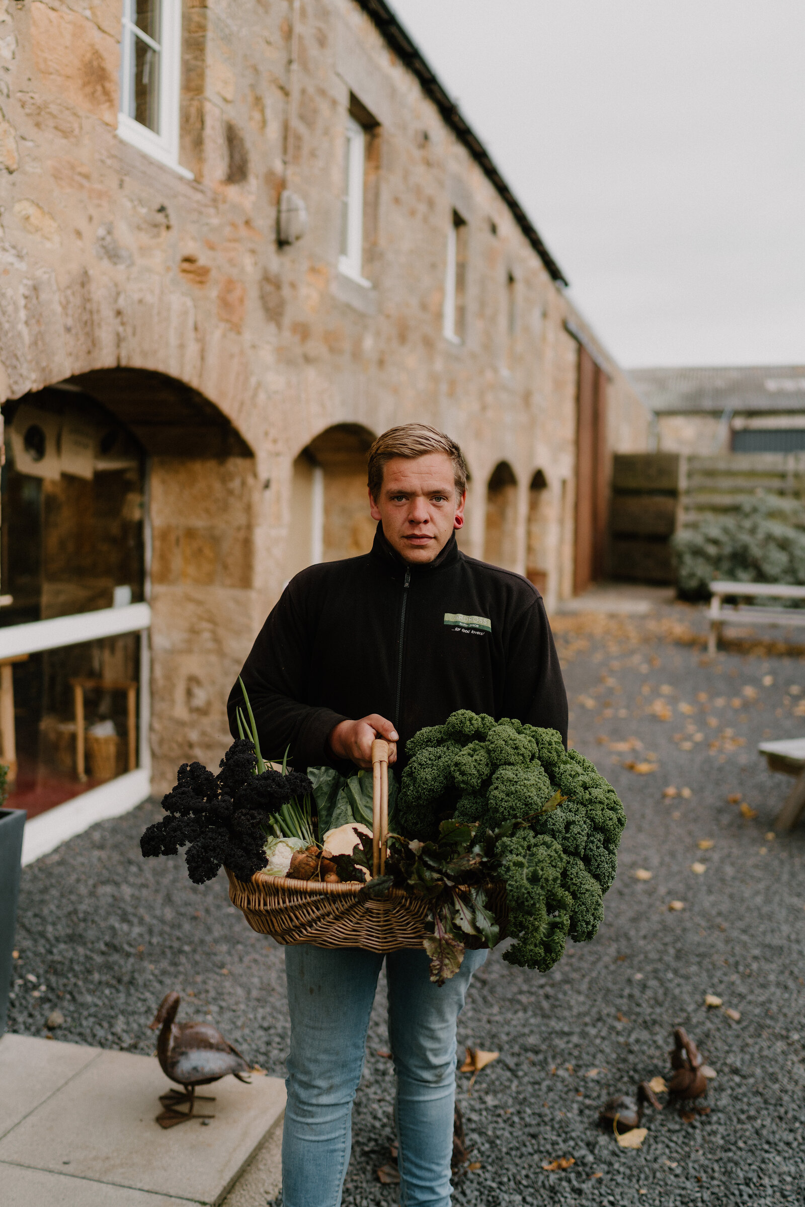 A man holds a wicker basket overflowing with kale, cauliflower and broccoli outside a stone farm building, duck ornaments standing on the gravel beside him.