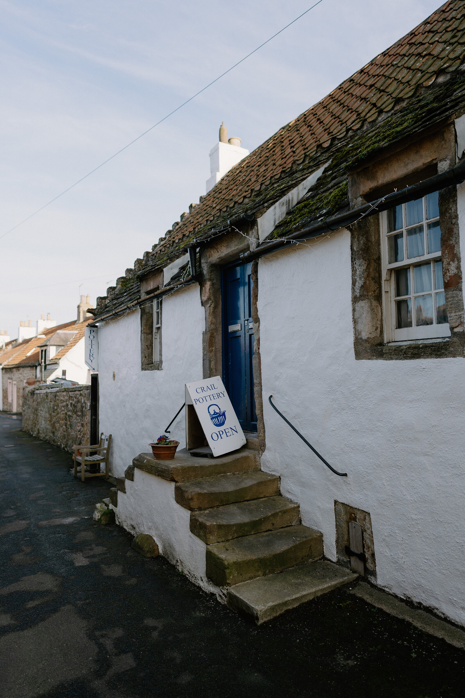 A sign reading "Crail Pottery, open" stands on stone steps beside a potted plant outside a whitewashed cottage with a pantile roof.