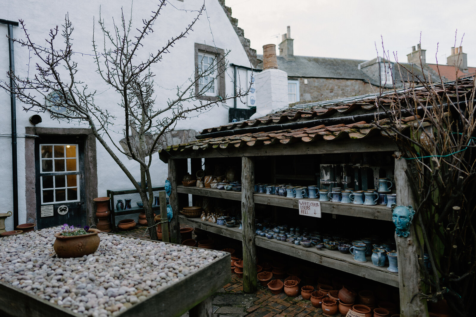 A wooden lean-to shelters rows of glazed pottery on shelves beside a stone cottage, a table of empty pots standing in the courtyard.
