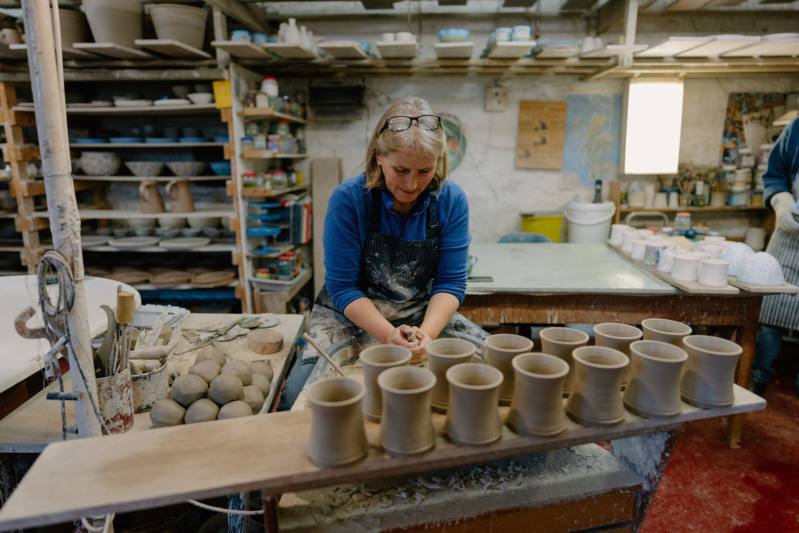 A potter in an apron trims a row of unfired mugs lined up on a wooden board in a workshop crowded with shelving.