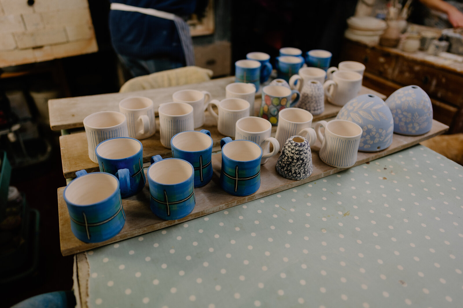 Rows of blue and white glazed mugs stand on a wooden board on a polka-dot tablecloth in a pottery workshop.