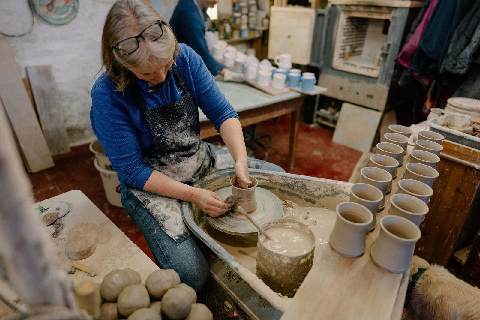 A woman in glasses and an apron throws a pot on a wheel, rows of finished mugs standing on a shelf beside her in a workshop.