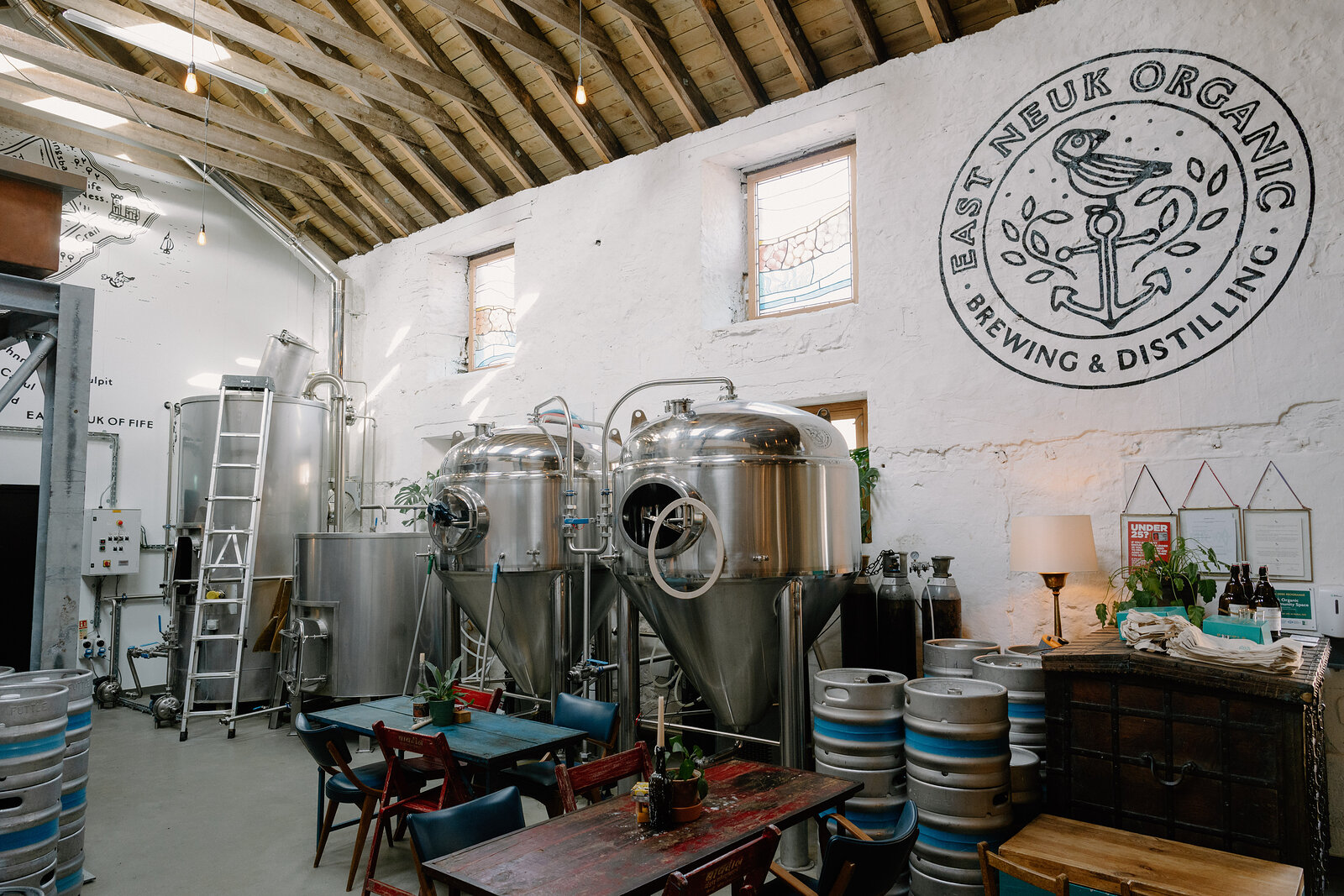 A roundel logo reading "EAST NEUK ORGANIC BREWING & DISTILLING" is painted on a whitewashed wall above steel brewing vessels, kegs and tables set out below.