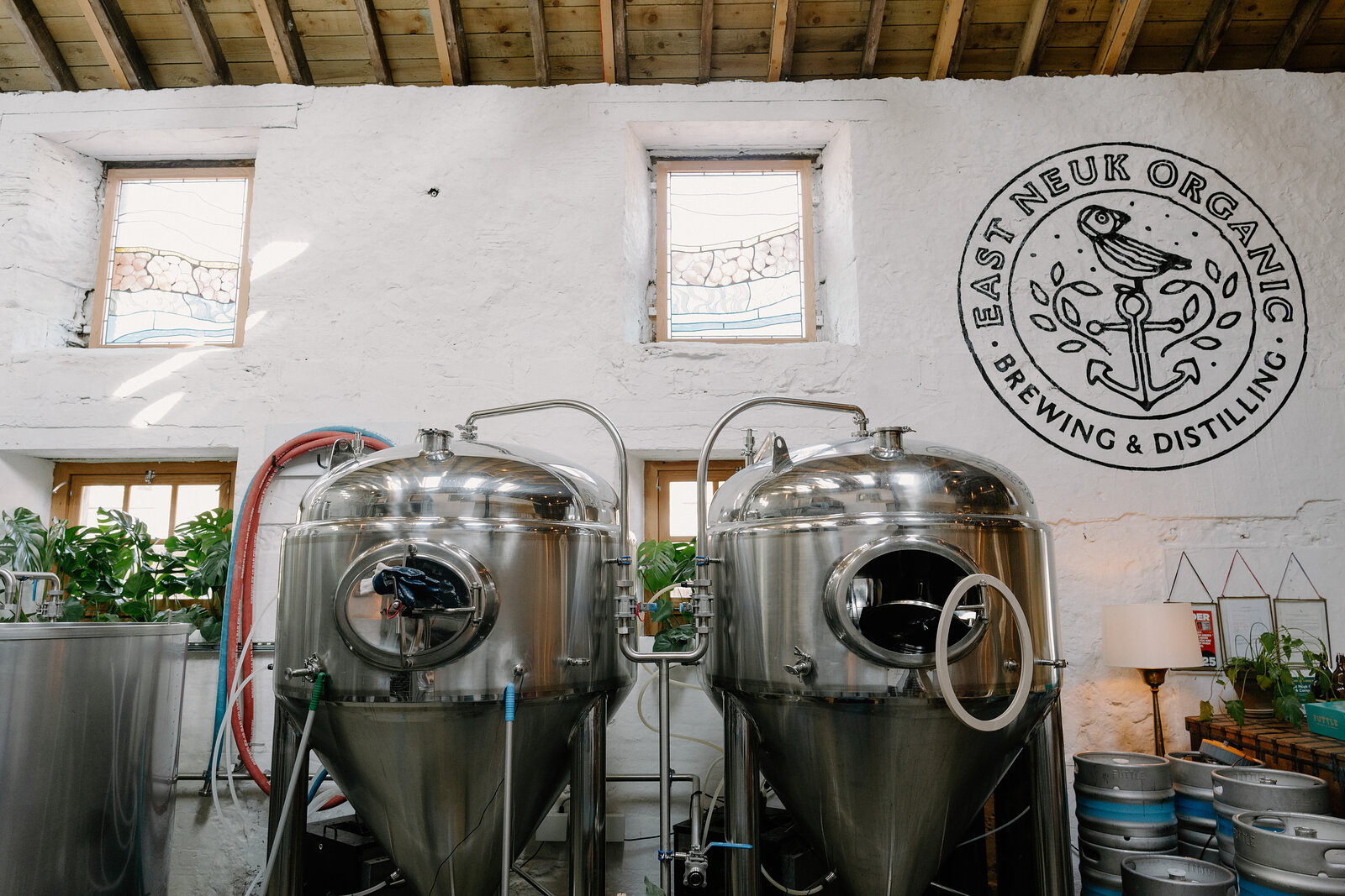 A roundel logo reading "EAST NEUK ORGANIC BREWING & DISTILLING" hangs above two steel fermentation tanks, houseplants and stacked kegs filling the room below.