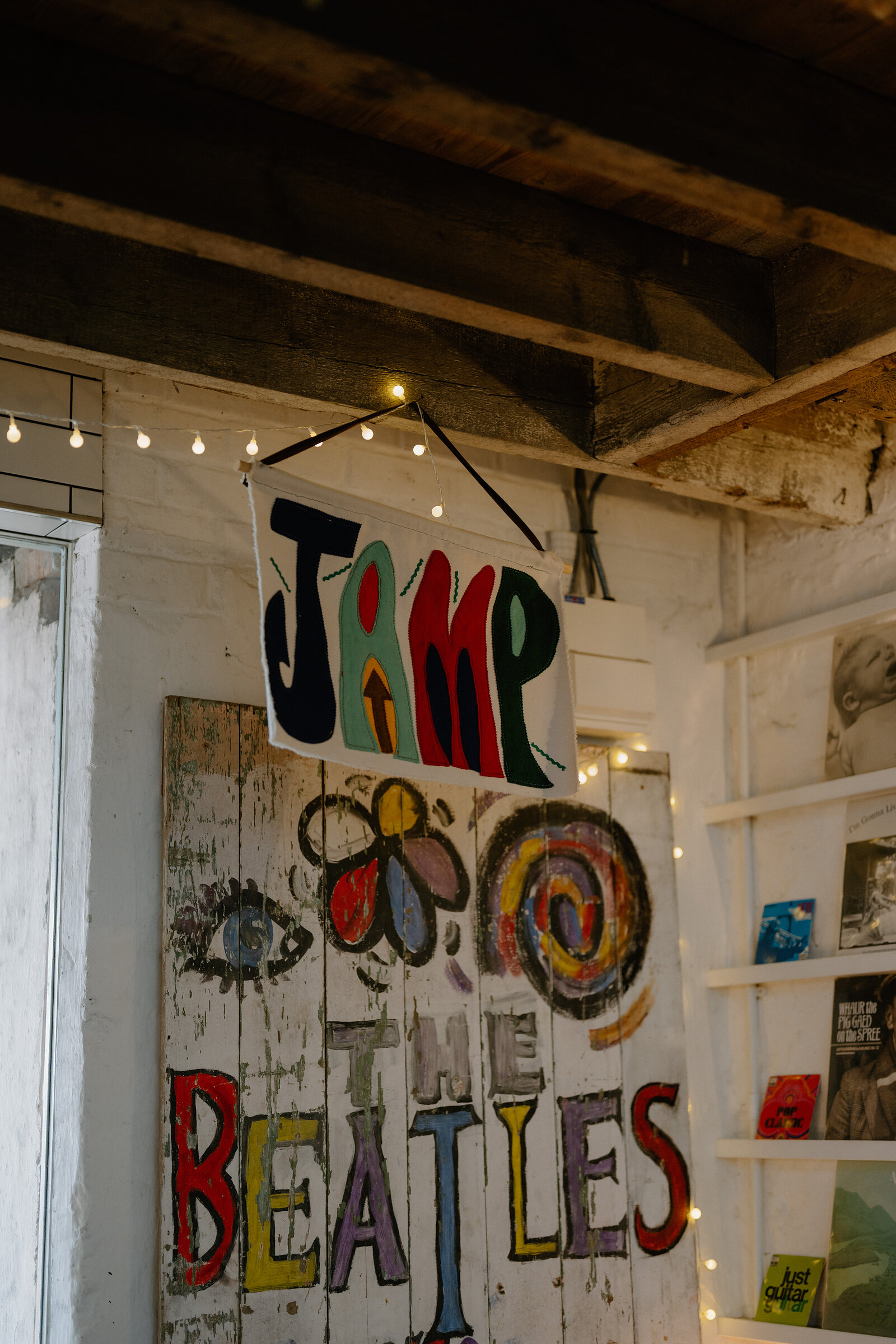 A hanging sign reading "JAMP" hangs above a colourful mural of a flower, a target and the words "THE BEATLES" painted on a weathered wooden door, record sleeves displayed on shelving beside it.