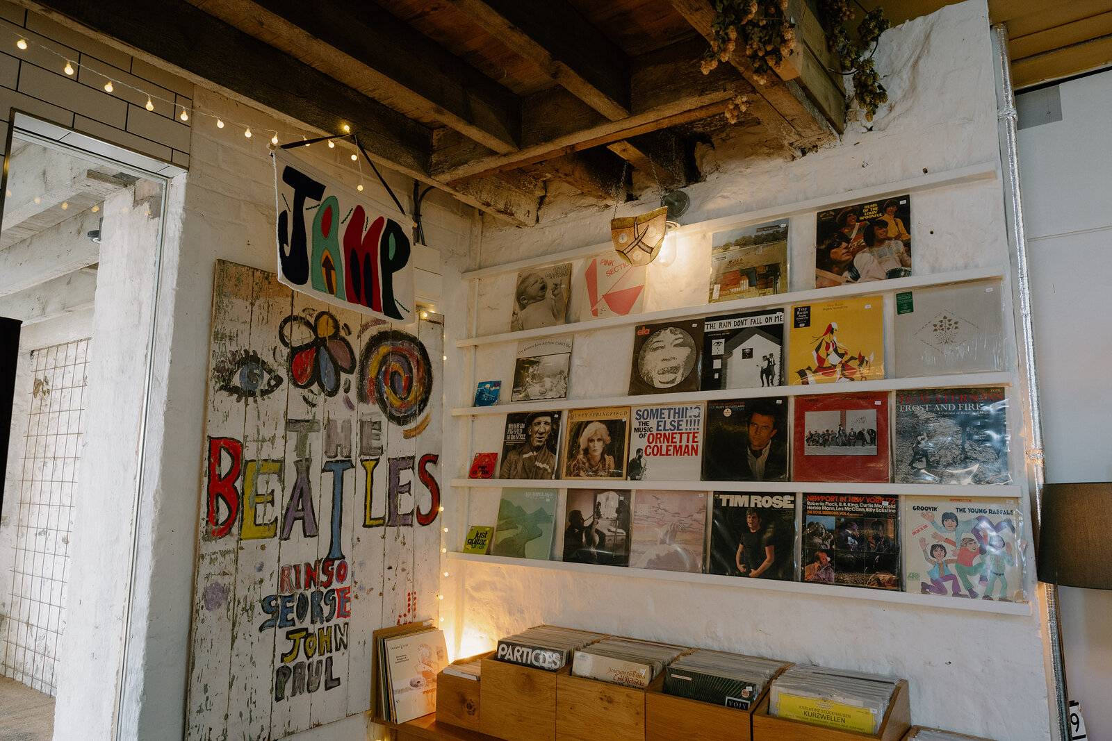 A mural reading "THE BEATLES" with the names "RINGO GEORGE JOHN PAUL" is painted on a door beside a wall hung with framed vinyl records and fairy lights.