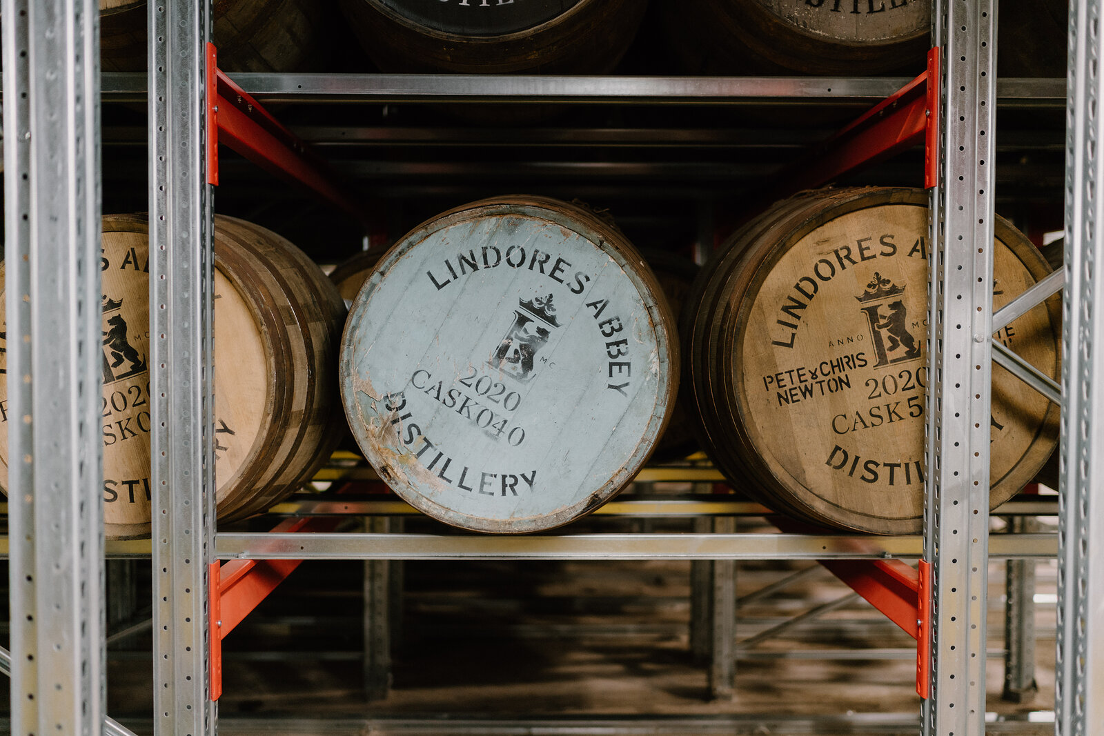 An oak whisky cask stamped 'Lindores Abbey Distillery, 2020, Cask 060' stands on steel racking beside other casks.