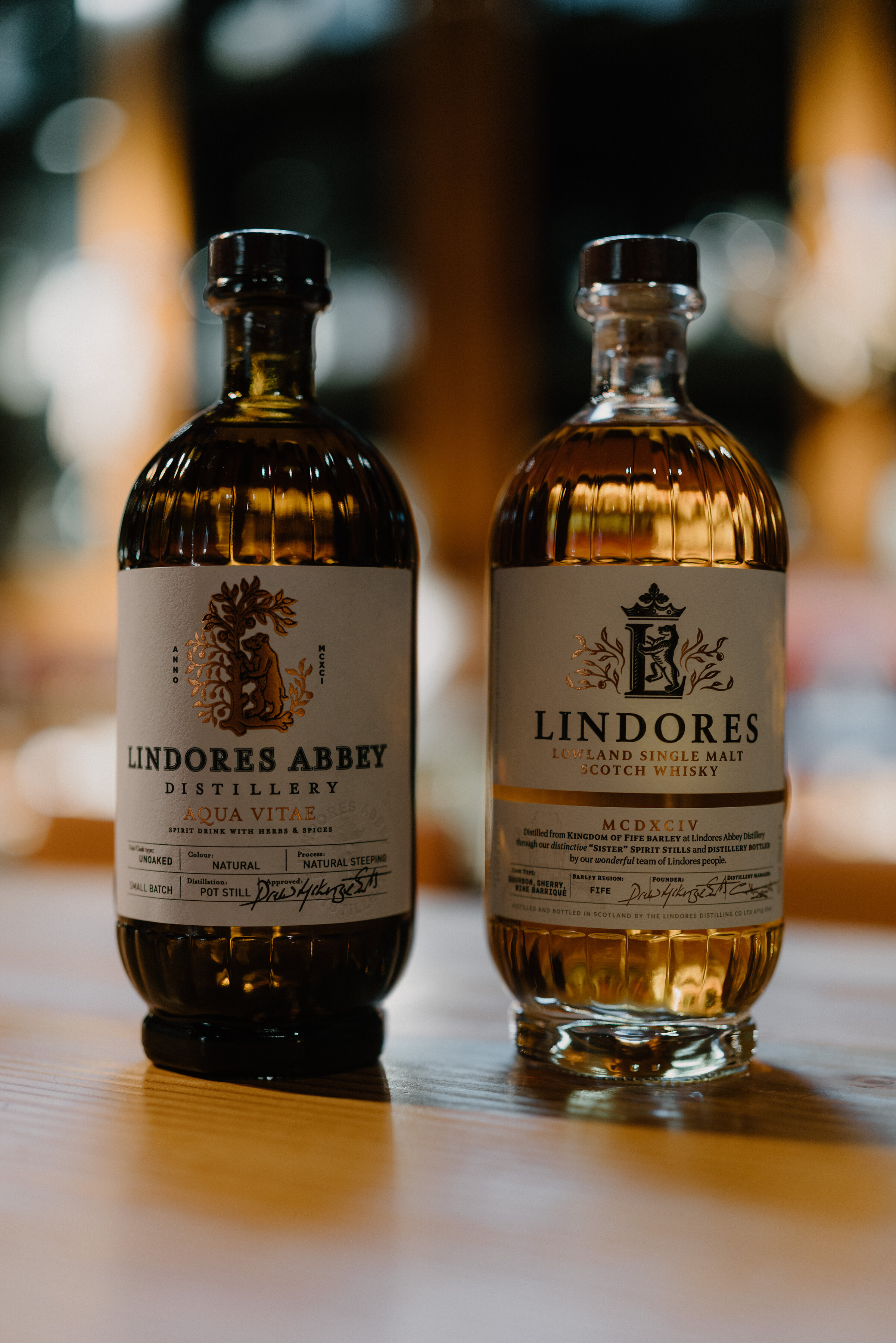 Two bottles of whisky, one labelled 'Lindores Abbey Distillery Aqua Vitae' and the other 'Lindores Lowland Single Malt Scotch Whisky', stand side by side on a wooden bar.