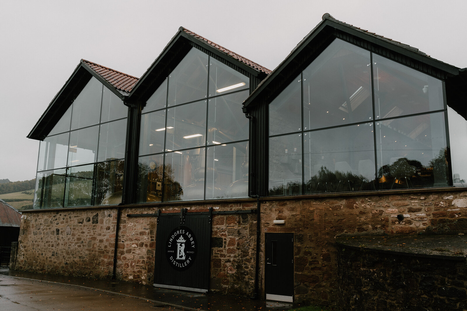 Three gabled glass extensions with black steel frames front a sandstone distillery building, a circular crest painted on a black door below.