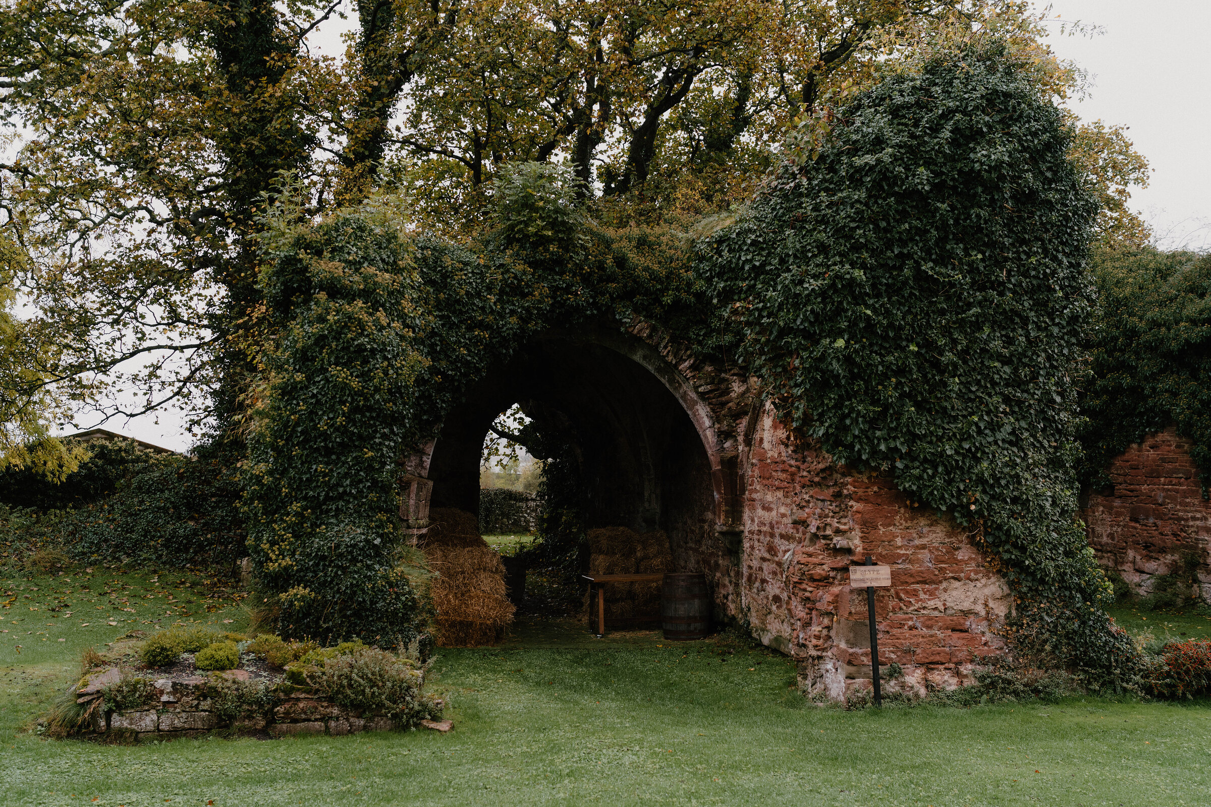 An ivy-covered stone archway stands in a ruined garden wall, a wooden bench visible through the opening on a lawn.