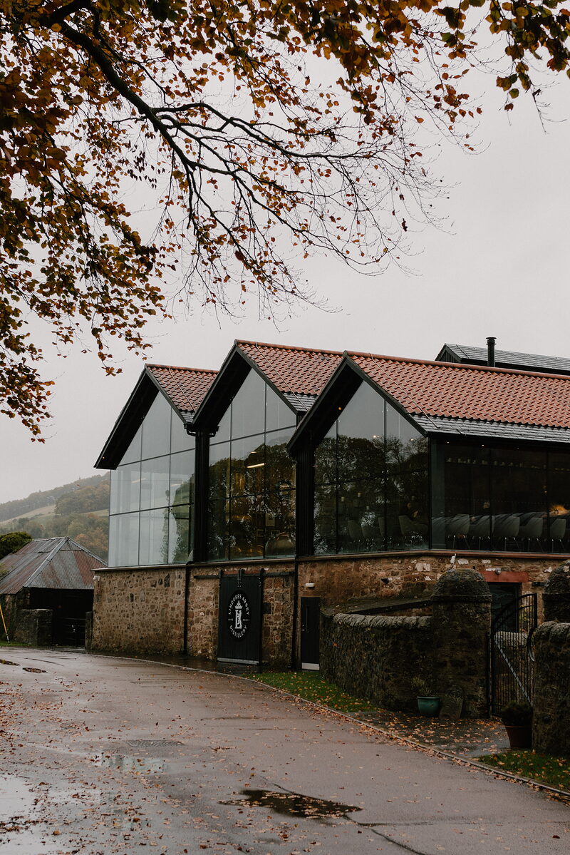 Bare autumn branches hang over a row of glass-gabled distillery buildings with a stone base, a wet lane running past a low stone wall.