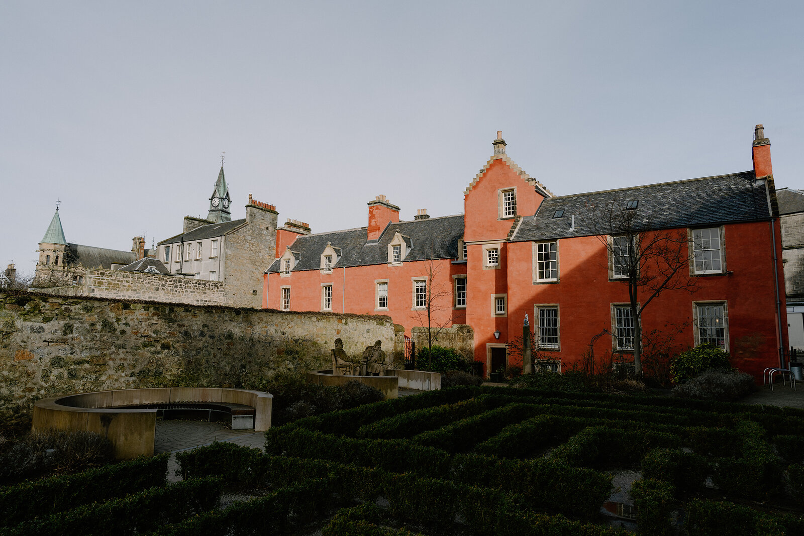 A salmon-pink painted house with stepped gables stands behind a clipped hedge maze, a castle wall and turret rising on the hillside behind.