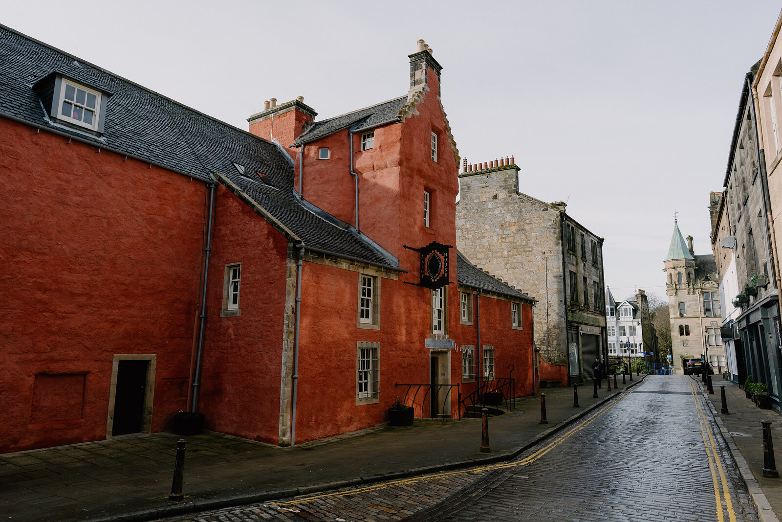 A red-painted historic building with stepped gables and chimneys stands on a cobbled street, a church tower rising in the distance.