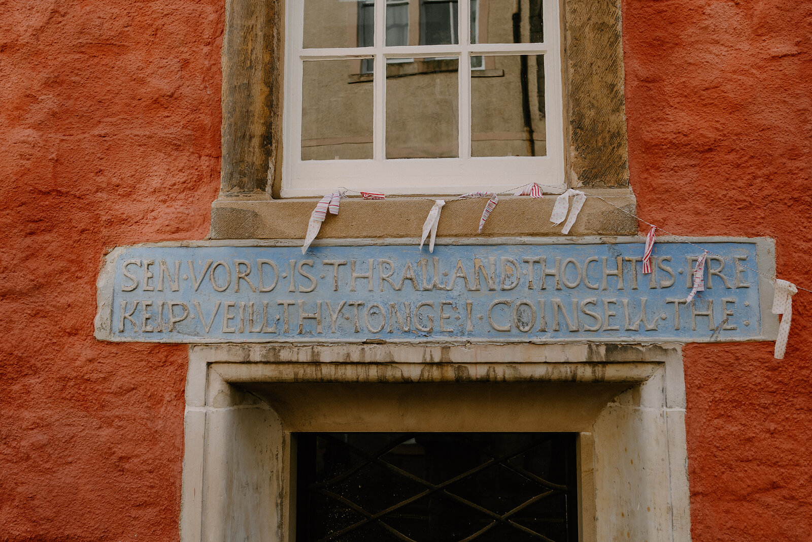 A weathered carved stone inscription in archaic lettering spans a lintel above a doorway on a red-painted building, a sash window above.