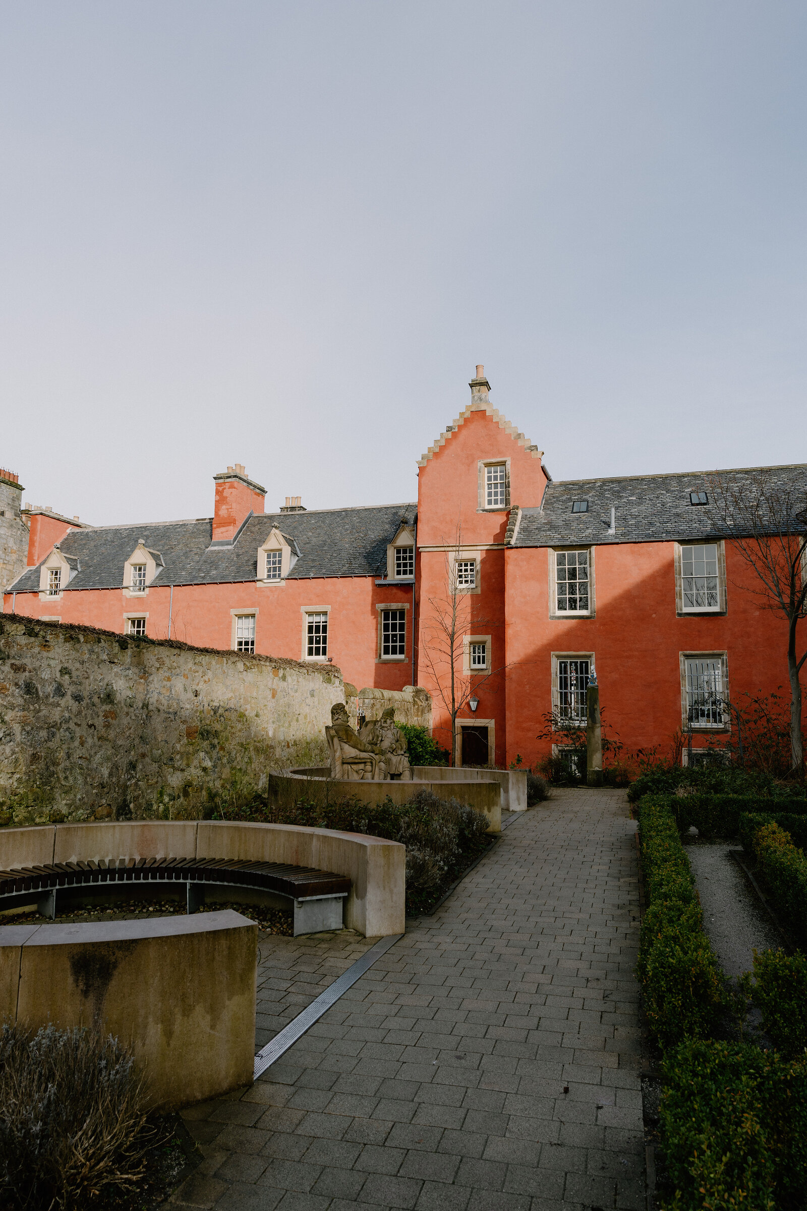 A salmon-pink painted house with stepped gables overlooks a walled garden with a stone pump and clipped hedges lining paved paths.