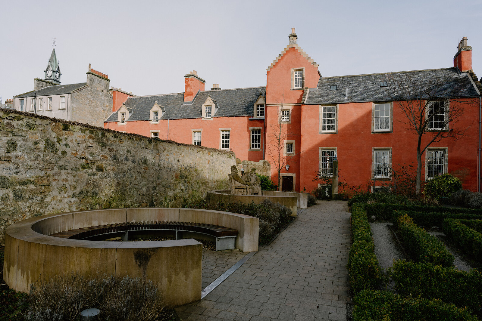A salmon-pink painted house with stepped gables overlooks a courtyard fountain and clipped hedges lining paved paths.