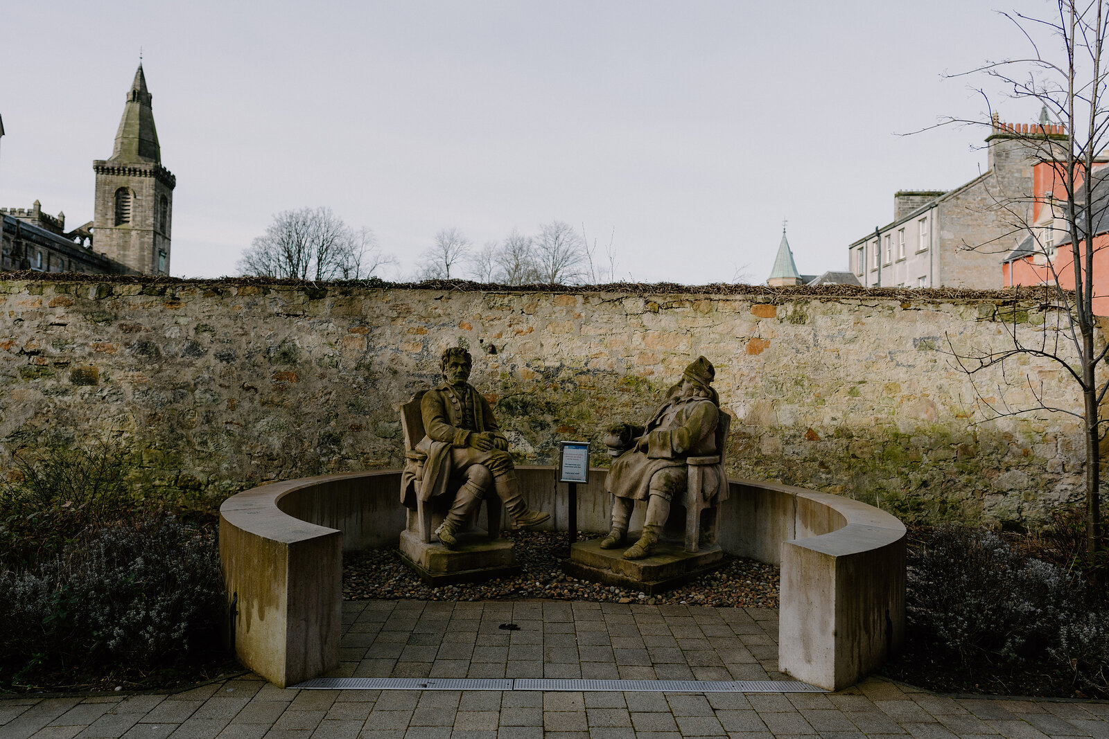Two bronze statues of seated figures rest on a curved stone bench before an old rubble wall, a church spire rising behind.