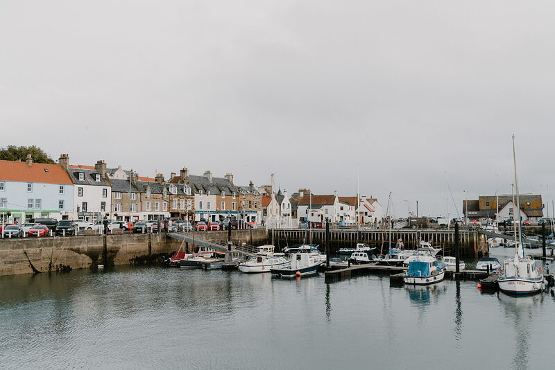 Boats are moored in a stone harbour beside a row of waterfront houses and shopfronts under a grey sky.