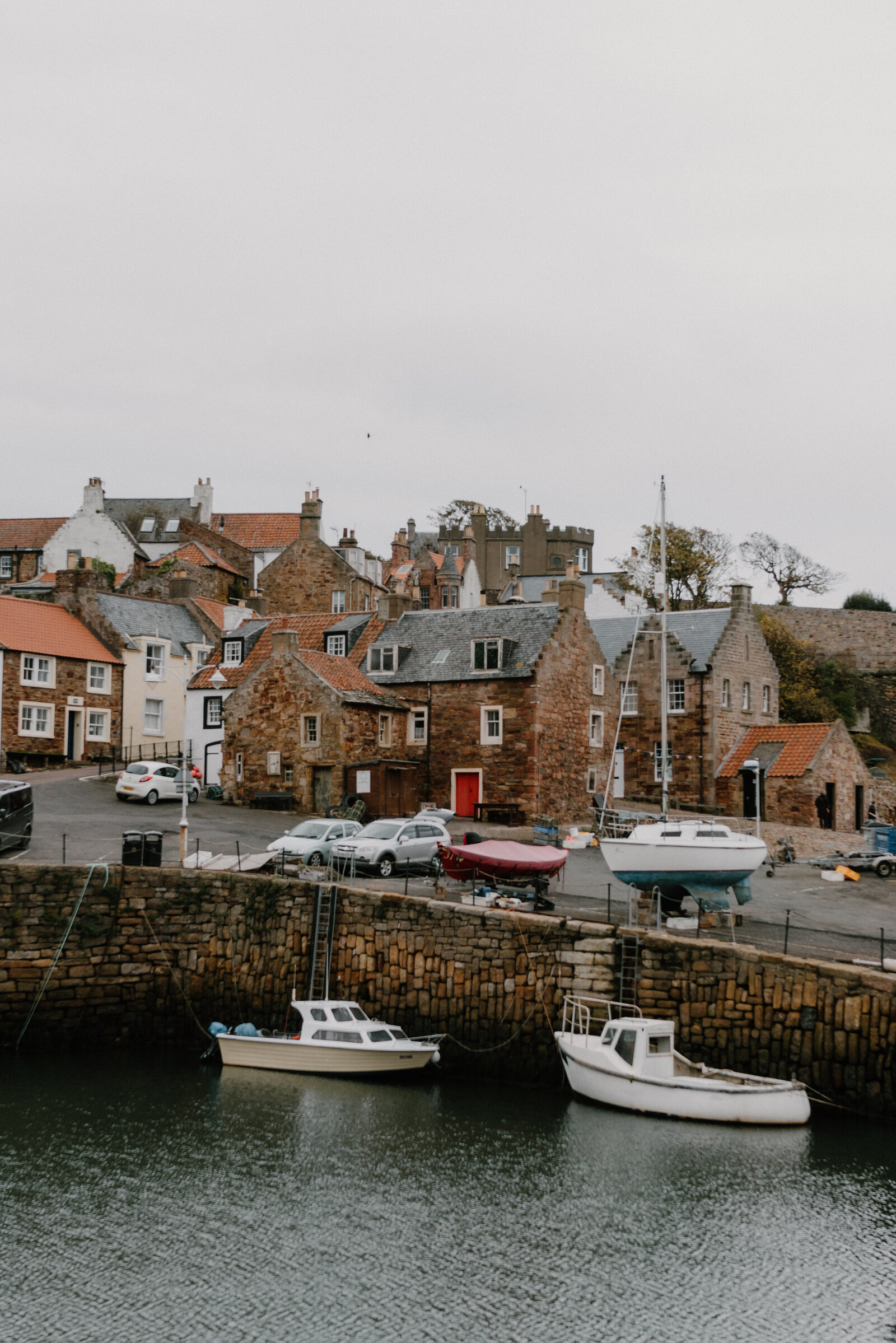 Fishing boats are moored in a stone harbour below a village of white and stone houses, cars parked along the quayside.