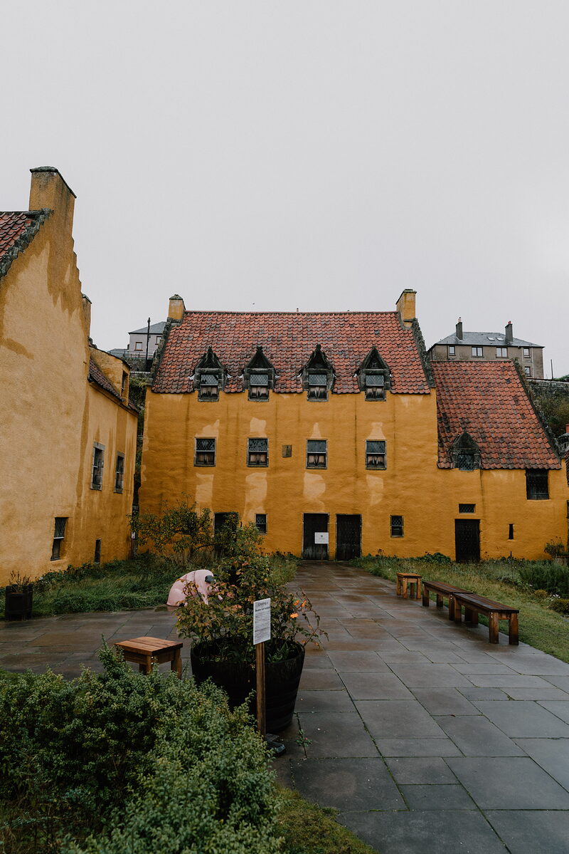 A large ochre-painted building with dormer windows and pantile roofs stands beyond a paved courtyard with potted plants and benches.
