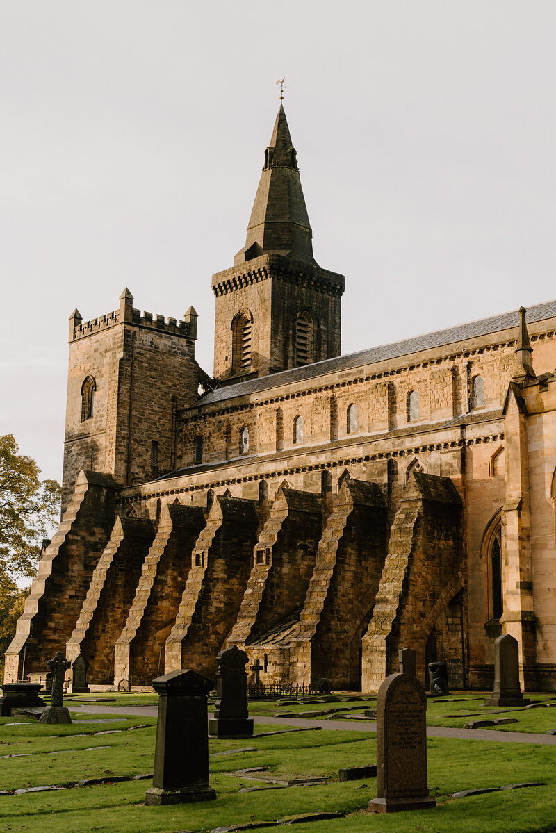 Stepped stone buttresses line the flank of a Gothic abbey with a central tower and spire, gravestones filling the churchyard below.